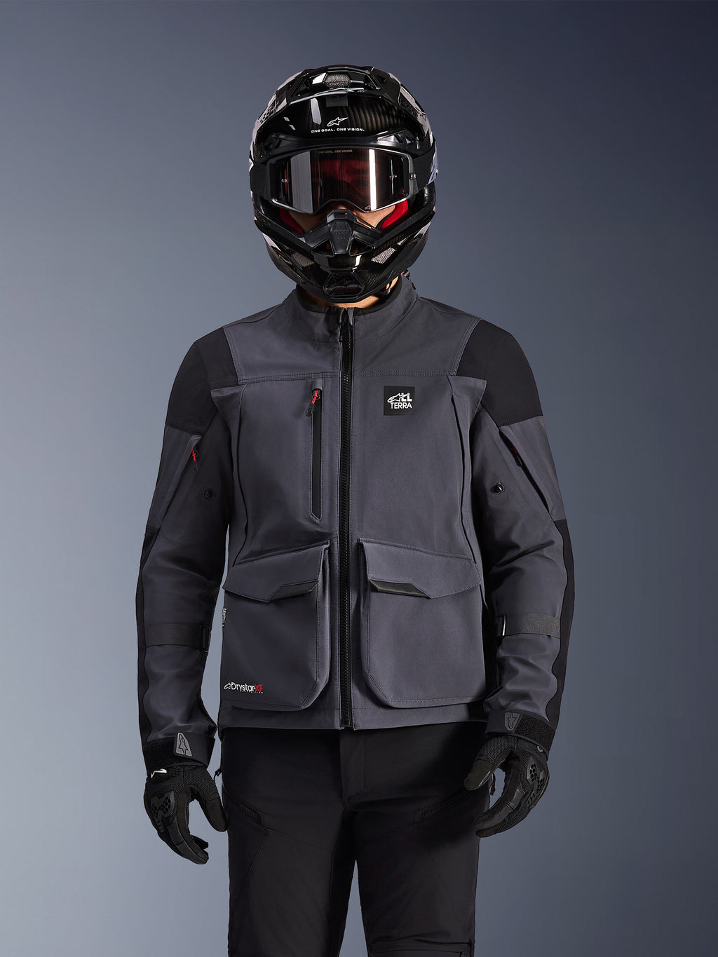 Alpinestars Maxdura Drystar®XF Laminated Armored Shell, motorjas, donkergrijs en zwart, gedragen door een rijder met een zwarte helm, bril en handschoenen, voorzien van grote technische cargozakken en ALL TERRA-branding, adventure touring uitrusting