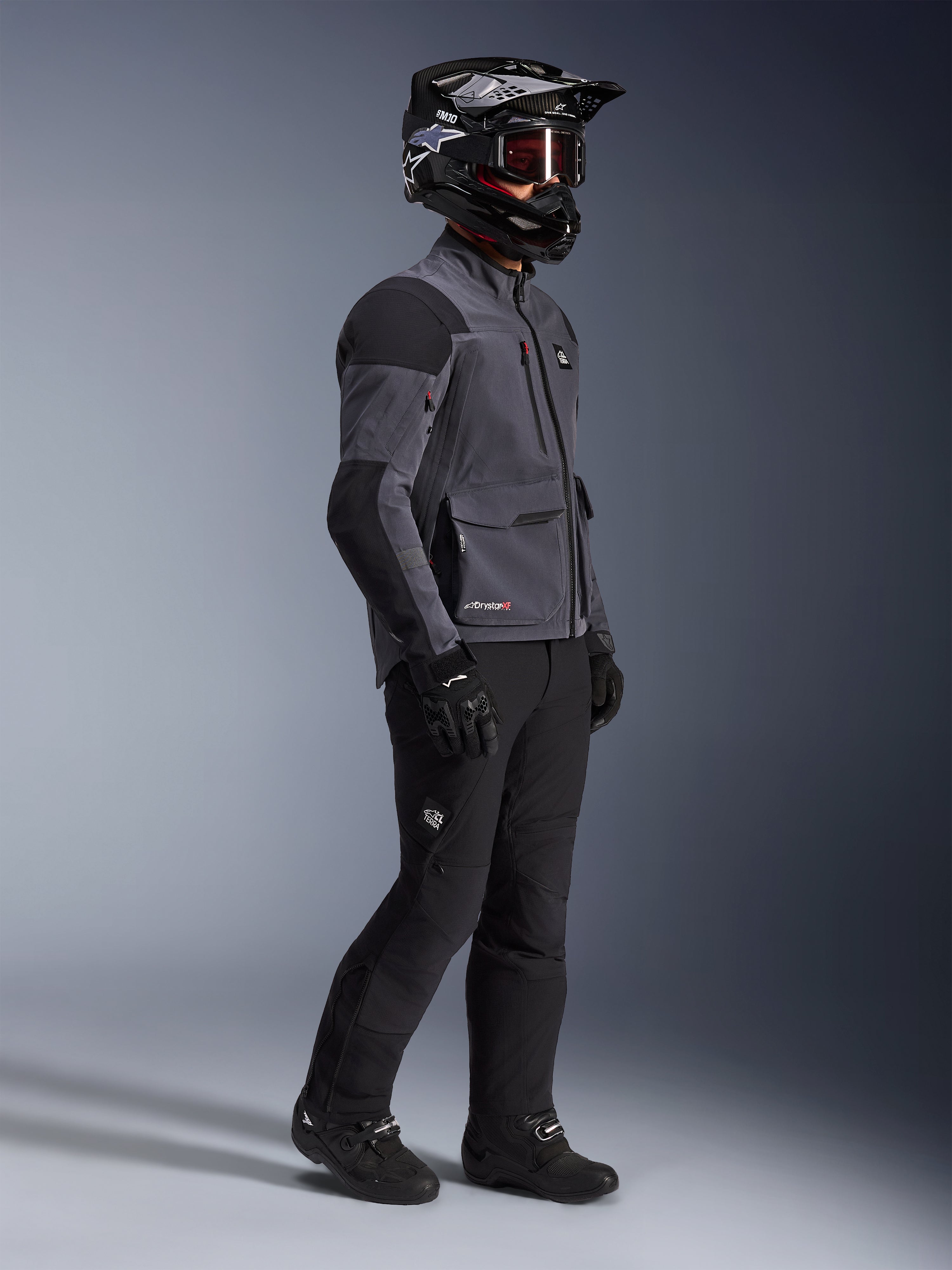 Motorrijder in Alpinestars Maxdura Drystar®XF Laminated Armored Shell jas in Deep Gray en Black, zwarte adventure motorbroek, zwarte helm met motorbril, zwarte handschoenen en laarzen, volledige studio-opname van de technische dual-sport uitrusting