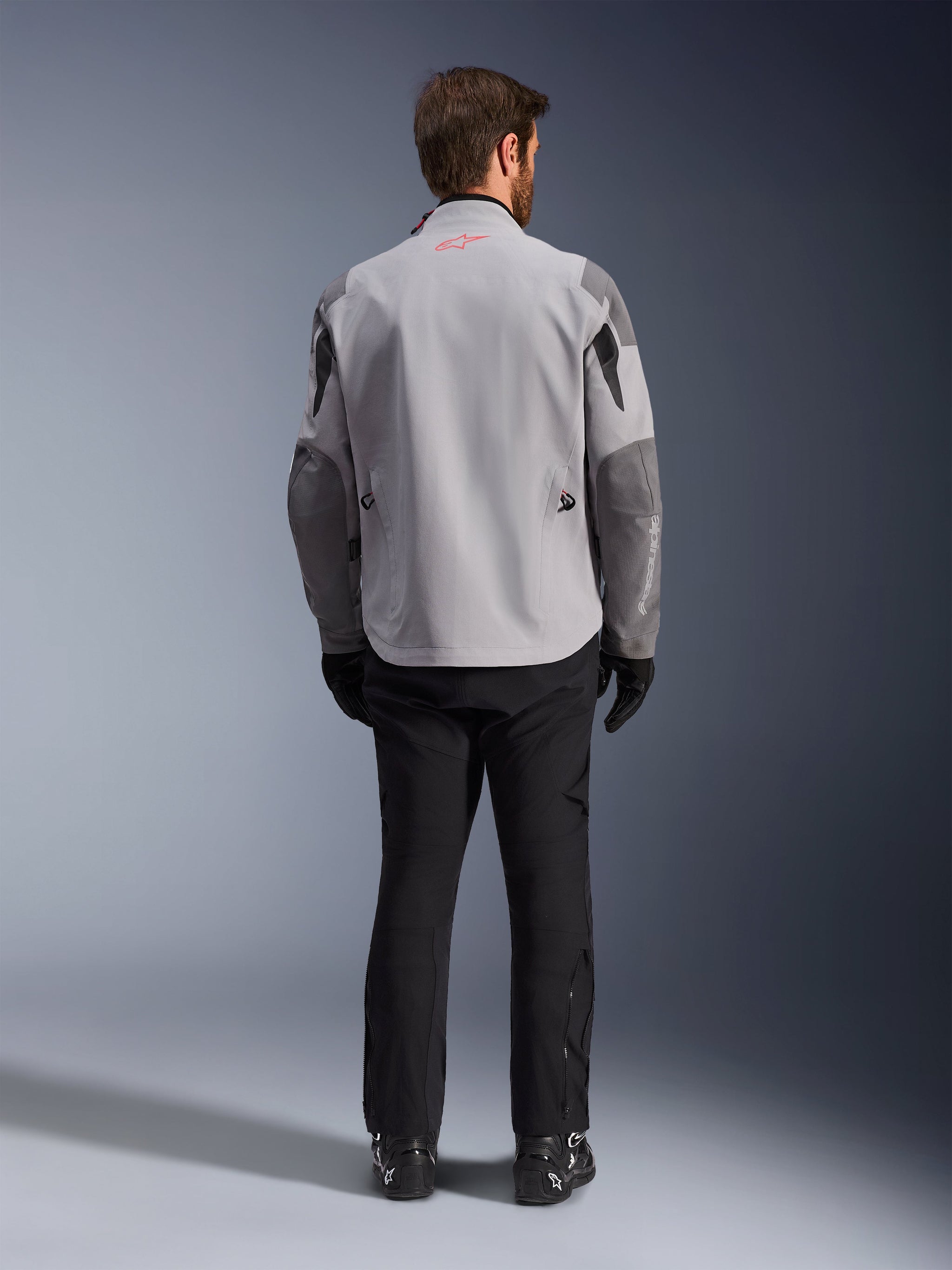 Alpinestars Maxdura Drystar®XF Laminated Armored Shell, Mid Gray and Dark Gray Motorcycle Jacket, back view, featuring a light gray body with dark gray panels on the shoulders and sleeves, small red Alpinestars star logo on the back collar, and white branding on the lower left sleeve, paired with black riding pants and boots.