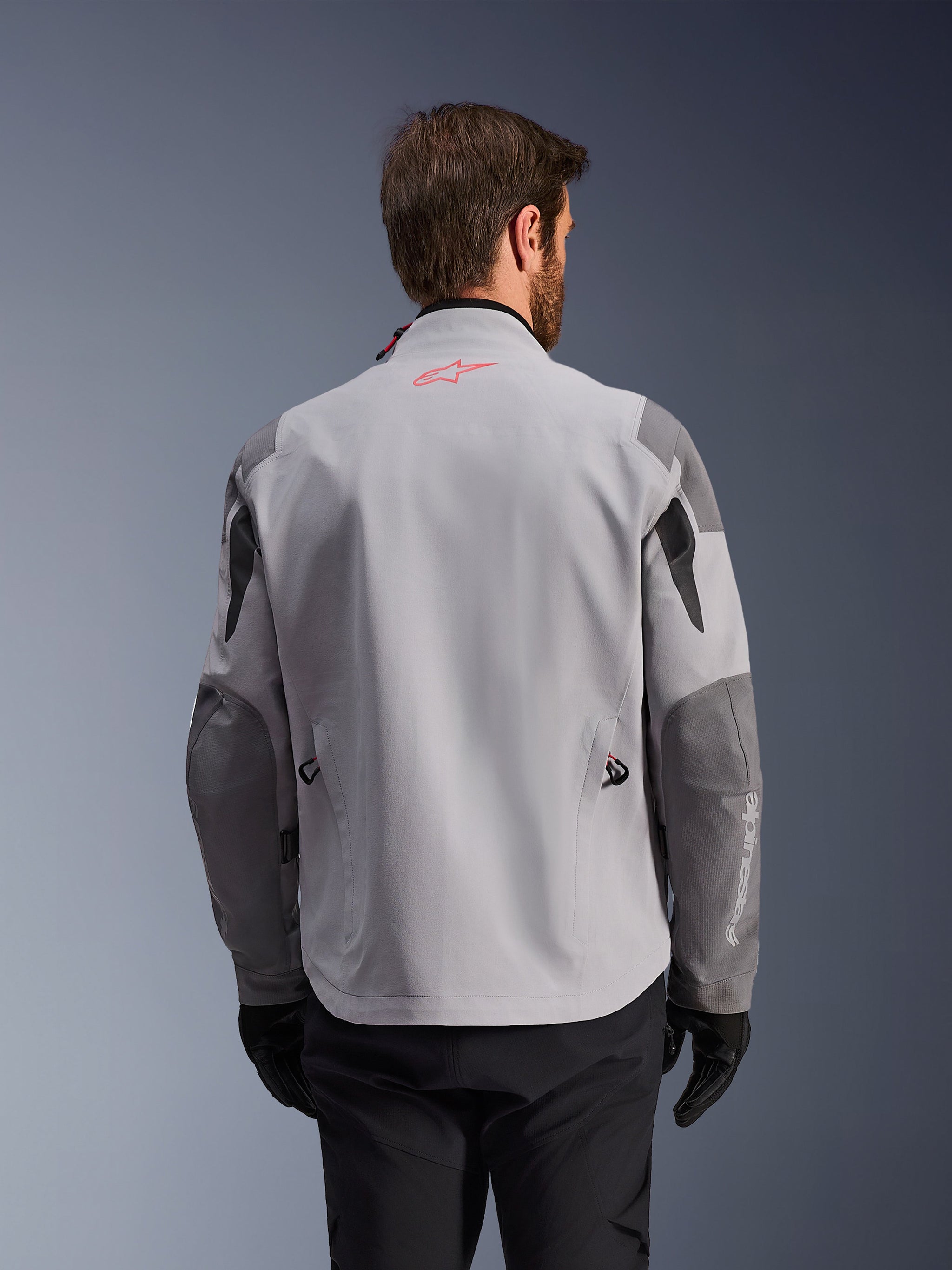 Alpinestars Maxdura Drystar®XF Laminated Armored Shell, back view of adventure motorcycle jacket, Mid Gray and Dark Gray, featuring a red star logo on the collar and white vertical branding on the sleeve, worn with black riding gloves and pants