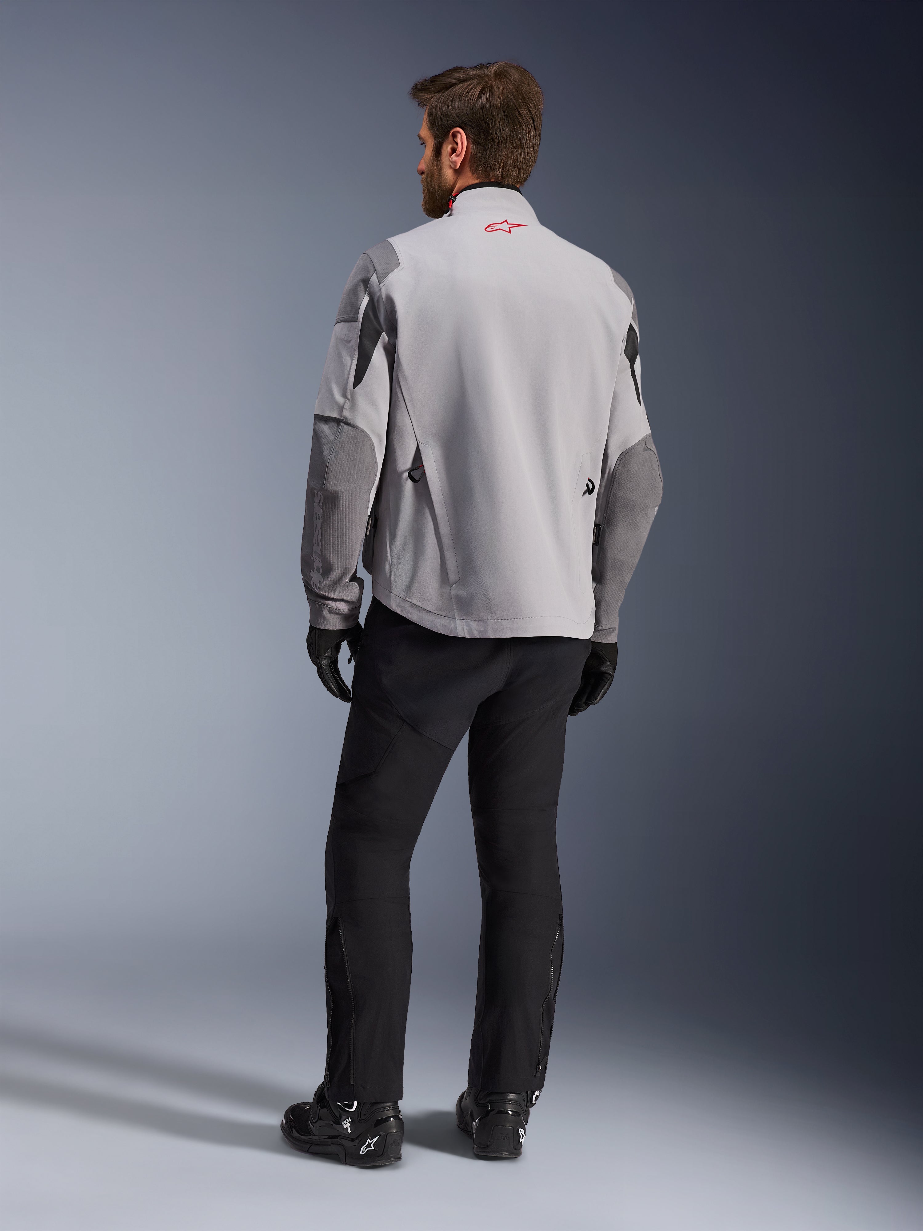 Maxdura Drystar®XF Gelamineerde Gepantserde Shell, motorjas, middengrijs en donkergrijs, achteraanzicht met rood Alpinestars-logo op de kraag, donkergrijze mouwverstevigingen, gedragen met zwarte broek en laarzen, adventure motorkleding