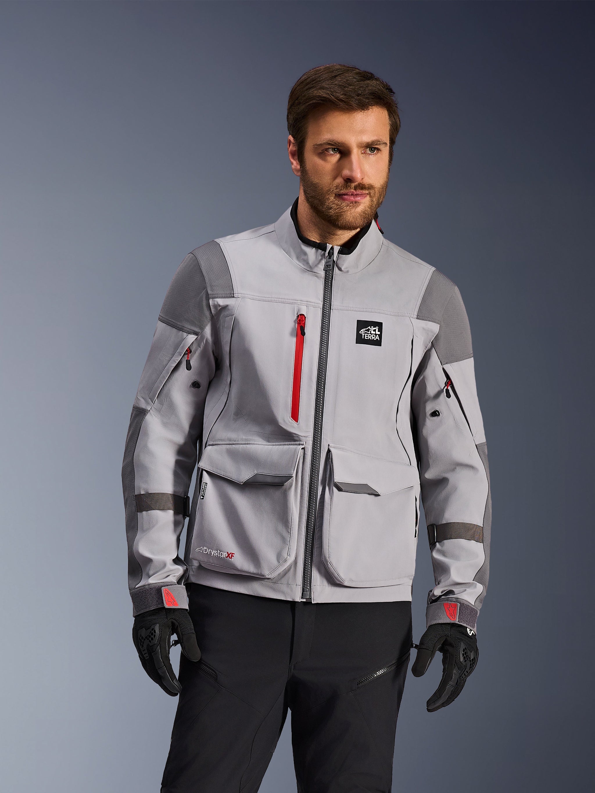 Alpinestars Maxdura Drystar®XF Laminated Armored Shell, Adventure Motorcycle Jacket, Mid Gray and Dark Gray with red accents, worn by a model with black gloves and pants, featuring large cargo pockets and waterproof zippers, professional riding apparel