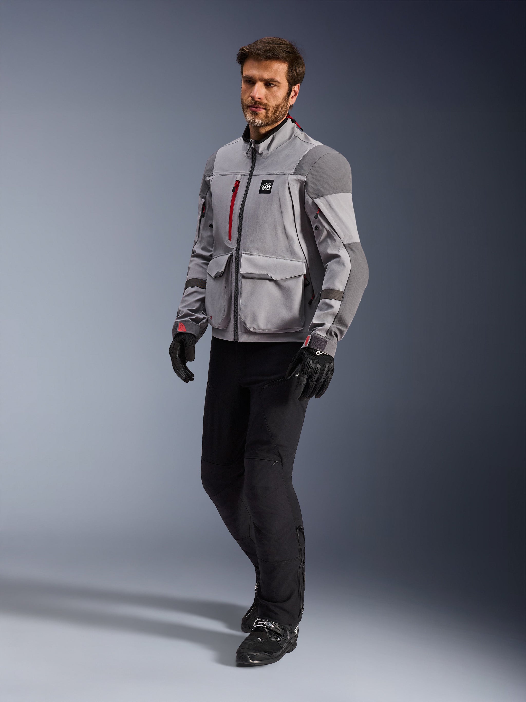 Alpinestars Maxdura Drystar®XF Laminated Armored Shell, Adventure Motorcycle Jacket, Mid Gray and Dark Gray, worn by a model with black riding pants and gloves, featuring red zipper accents and cargo pockets, professional all-weather technical gear for adventure touring