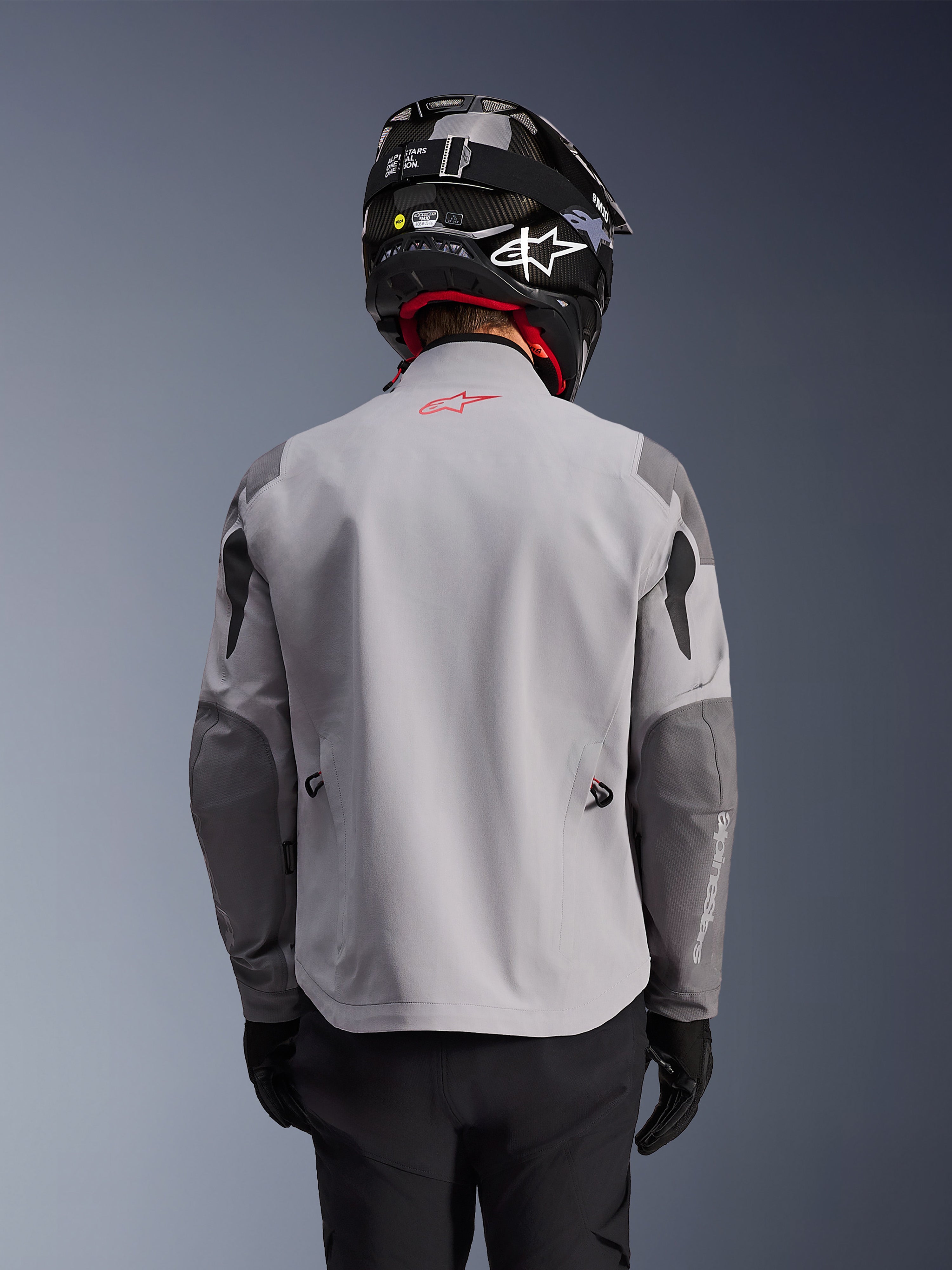 Alpinestars Maxdura Drystar®XF Gelamineerde Gepantserde Shell, adventure motorjas, middengrijs en donkergrijs, achteraanzicht met lichtgrijze body en donkergrijze versterkte panelen op schouders en ellebogen, rood Alpinestars-logo op de bovenrug, gedragen met een zwarte helm en zwarte handschoenen.