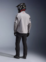 Rear view of a motorcycle rider wearing the Alpinestars Maxdura Drystar®XF Laminated Armored Shell jacket in Mid Gray and Dark Gray, featuring a red star logo on the collar, paired with black adventure pants, a black carbon-patterned helmet, and black boots.