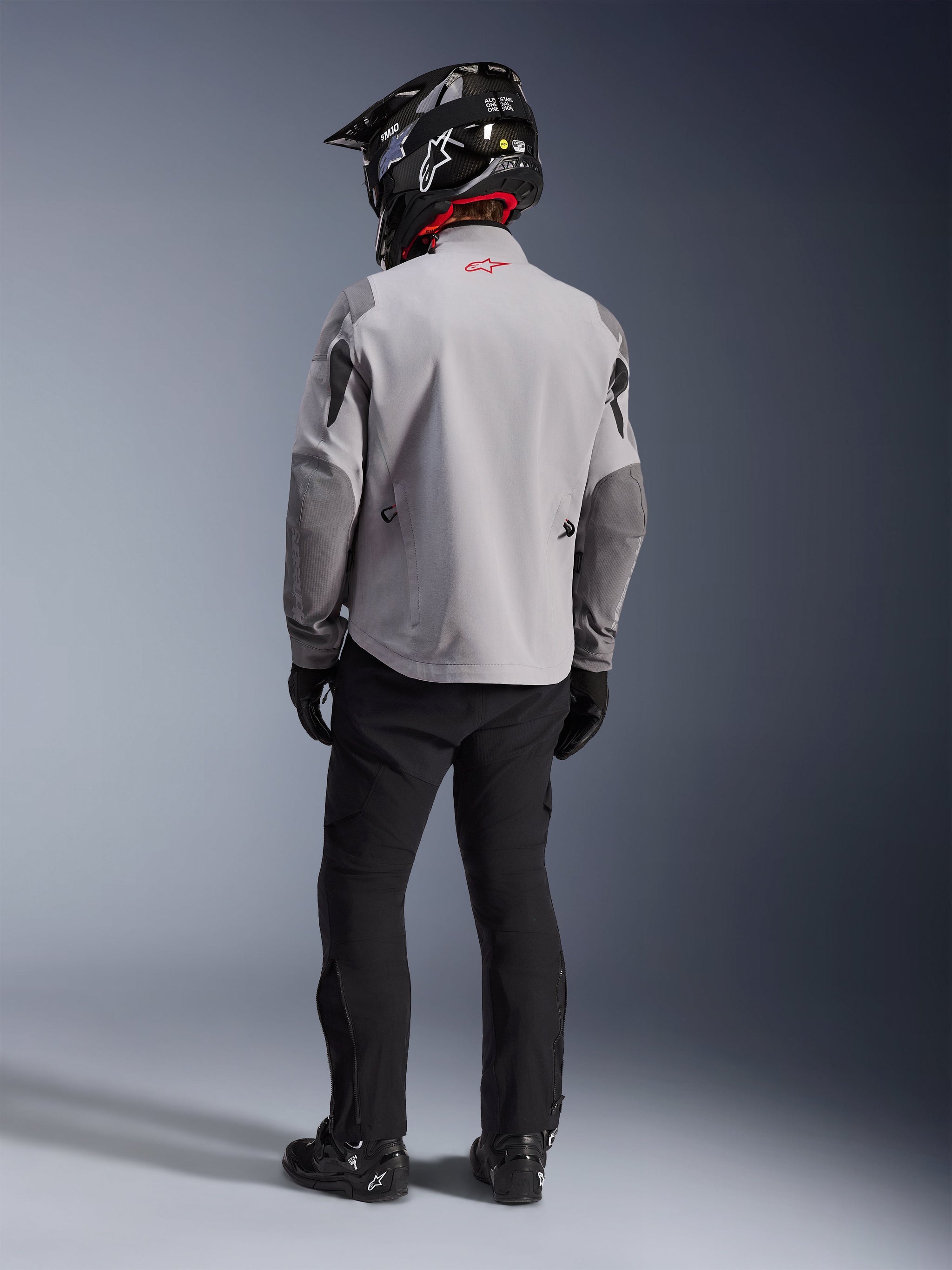 Rear view of a motorcycle rider wearing the Alpinestars Maxdura Drystar®XF Laminated Armored Shell jacket in Mid Gray and Dark Gray, featuring a red star logo on the collar, paired with black adventure pants, a black carbon-patterned helmet, and black boots.