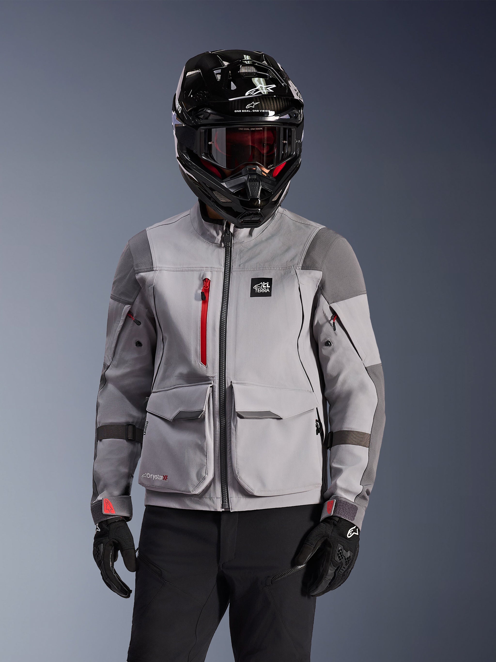 Alpinestars Maxdura Drystar®XF Laminated Armored Shell, adventure motorcycle jacket in Mid Gray and Dark Gray, worn with a black carbon-finish helmet and red-tinted goggles, featuring front cargo pockets and a red chest zipper, technical design for adventure touring.