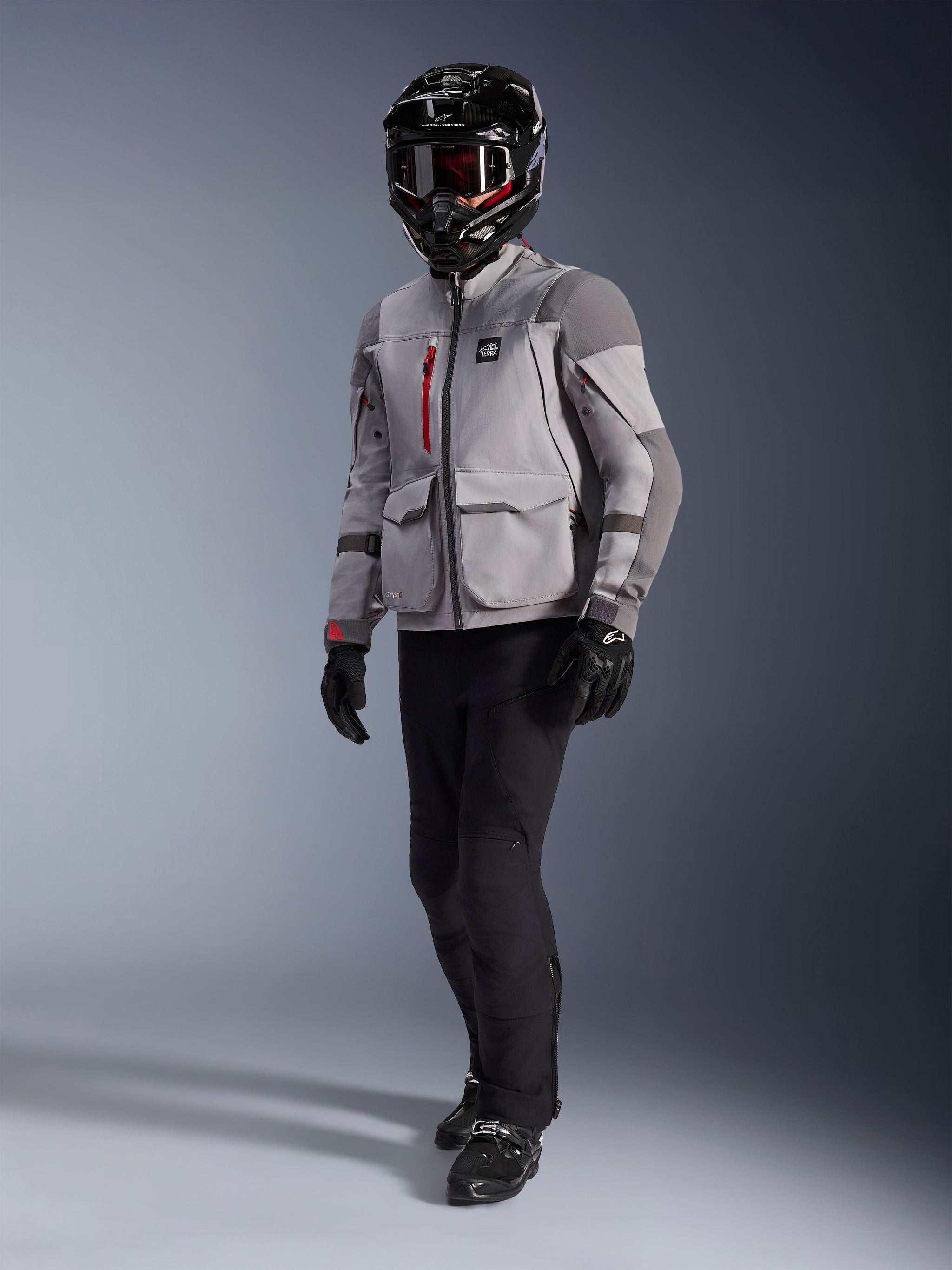 Adventure rider wearing the Alpinestars Maxdura Drystar®XF Laminated Armored Shell, Mid Gray and Dark Gray motorcycle jacket, black adventure helmet with goggles, black pants and gloves, featuring red zipper accents and cargo pockets, technical gear for adventure touring.