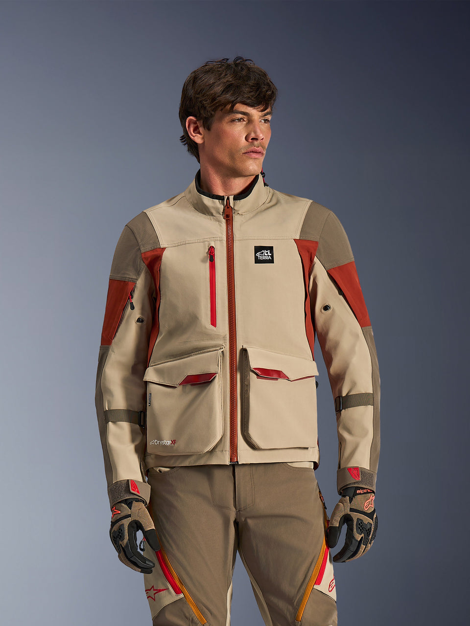 Alpinestars Maxdura Drystar®XF Laminated Armored Shell, Aluminum Walnut Rust Brown, adventure motorjas gedragen door een mannelijk model, voorzien van grote cargozakken aan de voorkant en technische rode accenten, gecombineerd met een bijpassende adventure motorbroek en handschoenen