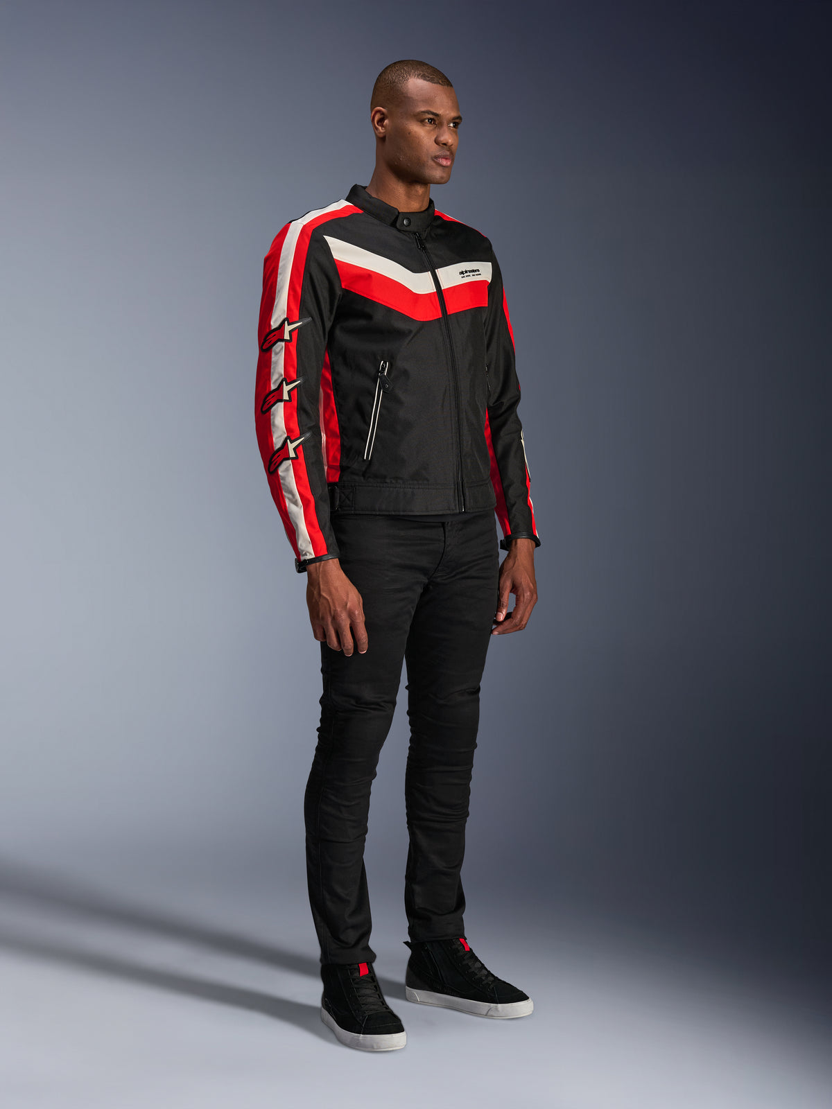Mannelijk model in Alpinestars T-Dyno WR Jas, textiele motorjas in Black Bright Red Ecru, met rode en gebroken witte strepen en Alpinestars sterlogo's op de mouwen, gecombineerd met een zwarte broek en zwarte hoge sneakers, vintage-geïnspireerde urban motorkleding.