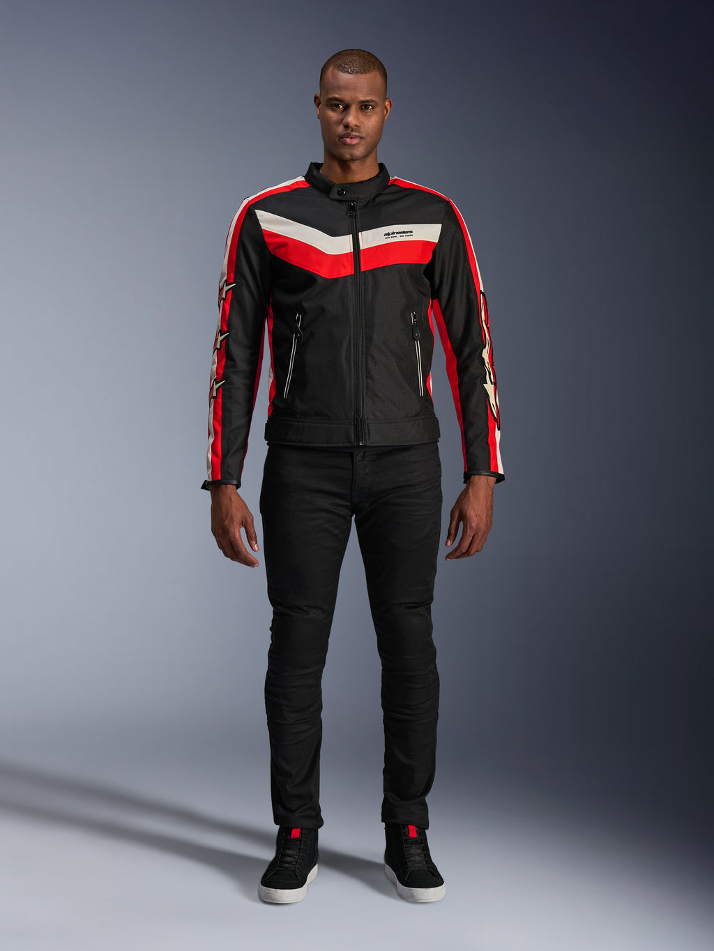 Alpinestars T-Dyno WR Jas, textiele motorjas in zwart, felrood en ecru, met V-vormige strepen op de borst en ster-logo's op de mouwen, gedragen met een zwarte broek en zwarte motorschoenen, vintage-geïnspireerde urban gear