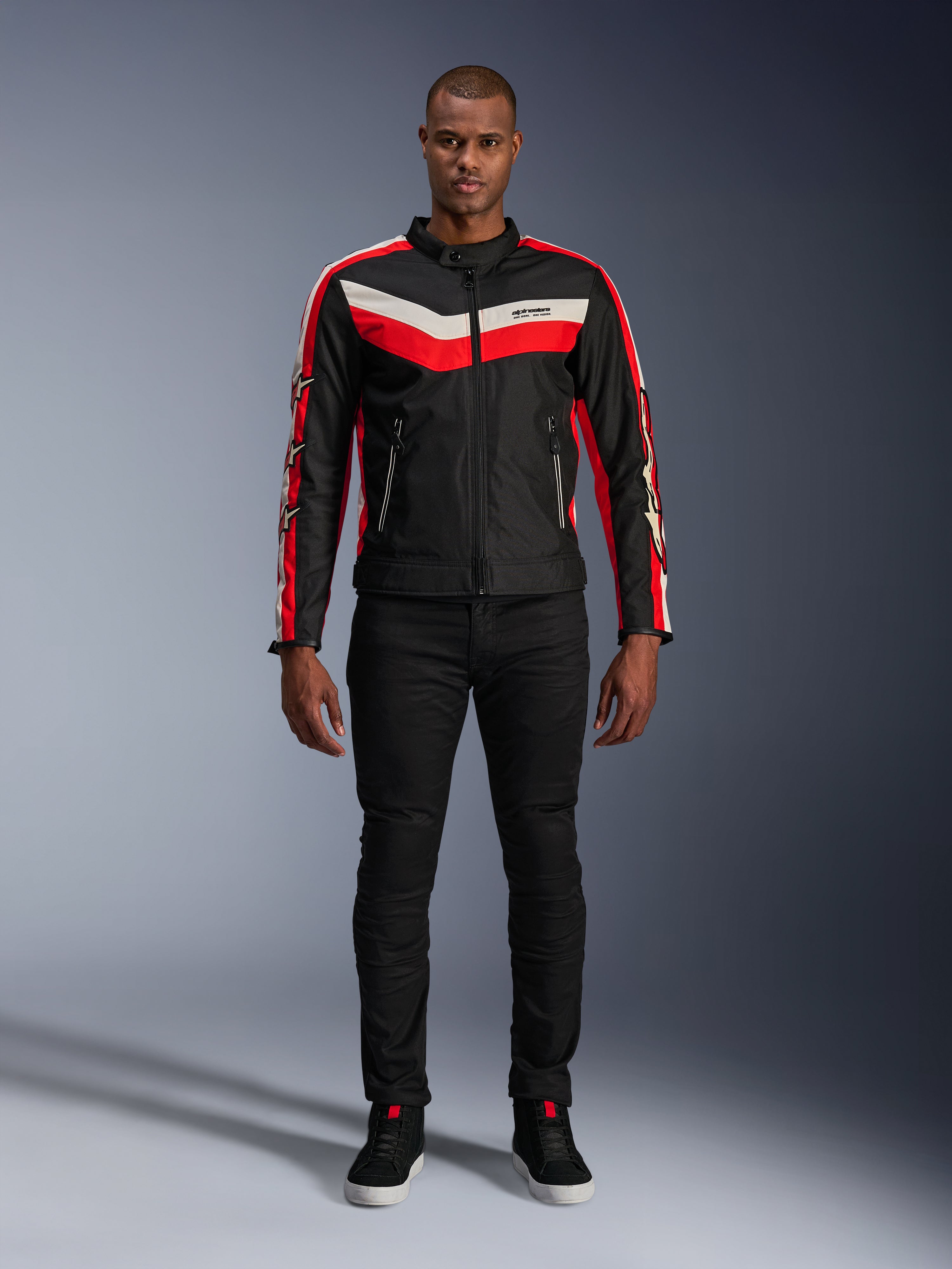 Alpinestars T-Dyno WR Jas, textiele motorjas in zwart, felrood en ecru, met V-vormige strepen op de borst en ster-logo's op de mouwen, gedragen met een zwarte broek en zwarte motorschoenen, vintage-geïnspireerde urban gear