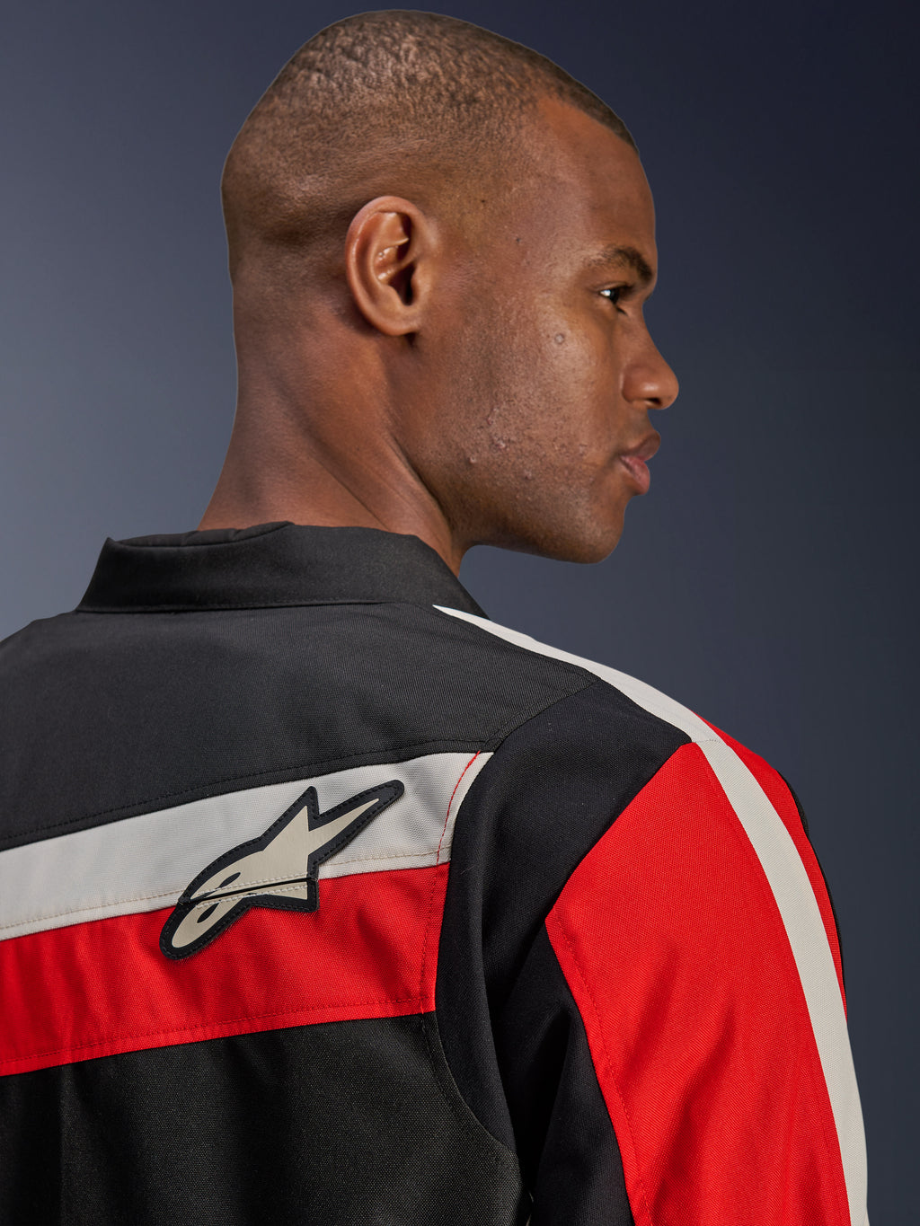 Alpinestars T-Dyno WR-jas, close-up achteraanzicht op een model, textiele motorjas in zwart met horizontale ecru en felrode strepen, voorzien van de Alpinestars ster-logopatch op de schouder, op erfgoed geïnspireerde bikerstijl