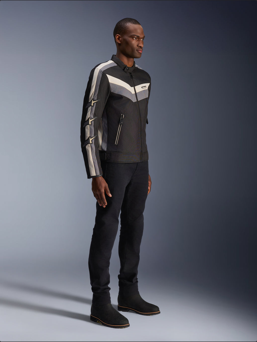 Alpinestars T-Dyno WR Jas, textiele motorjas, zwart met donkergrijze en ecru chevron-accenten, vintage-geïnspireerde bikerpasvorm, gedragen met zwarte broek en laarzen voor een complete road-riding look