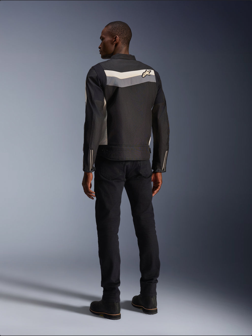 Mannelijk model van achteren gezien die de Alpinestars T-Dyno WR Jas in Black Dark Gray Ecru draagt, textiele motorjas met een horizontale streep en sterlogo op de rug, gecombineerd met een zwarte motorbroek en laarzen, urban motoruitrusting esthetiek.