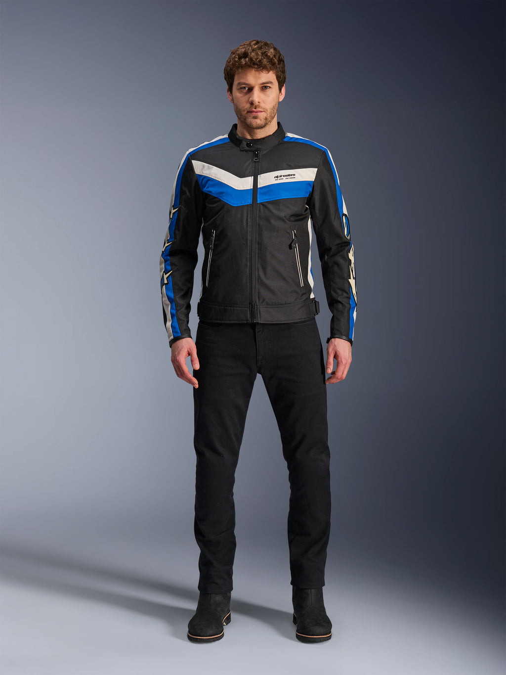 Alpinestars T-Dyno WR Jacket, textiele motorjas, zwart, blauw en ecru, gedragen door een mannelijk model met zwarte broek en laarzen, met chevron-strepen op de borst en erfgoed biker-ontwerp