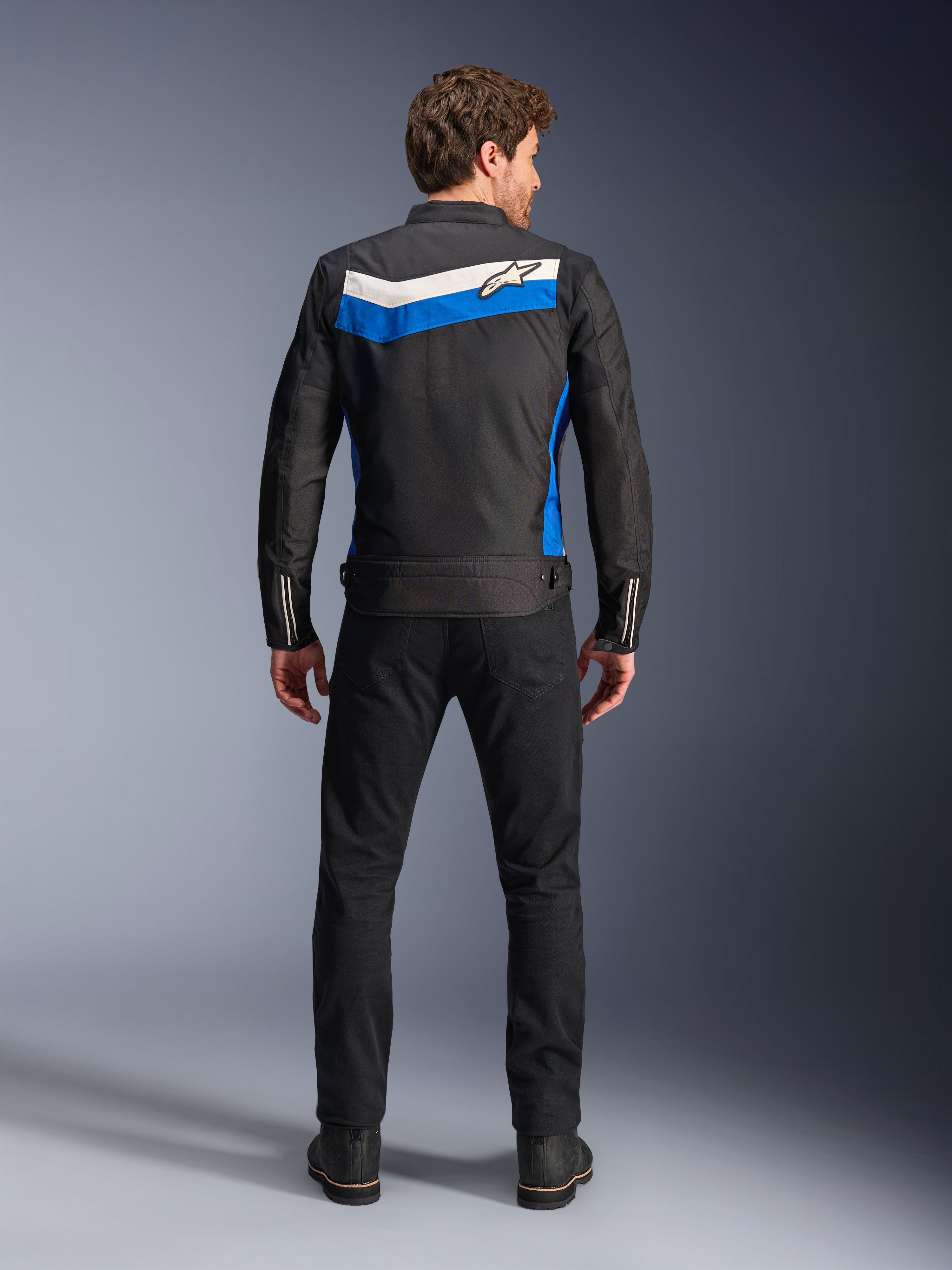 Alpinestars T-Dyno WR Jas, textiele motorjas, kleurstelling Black Blue Ecru, volledig achteraanzicht op model, voorzien van een blauwe en ecru chevron-streep met Alpinestars-logo, gecombineerd met zwarte broek en laarzen, vintage-geïnspireerde urban motorkleding