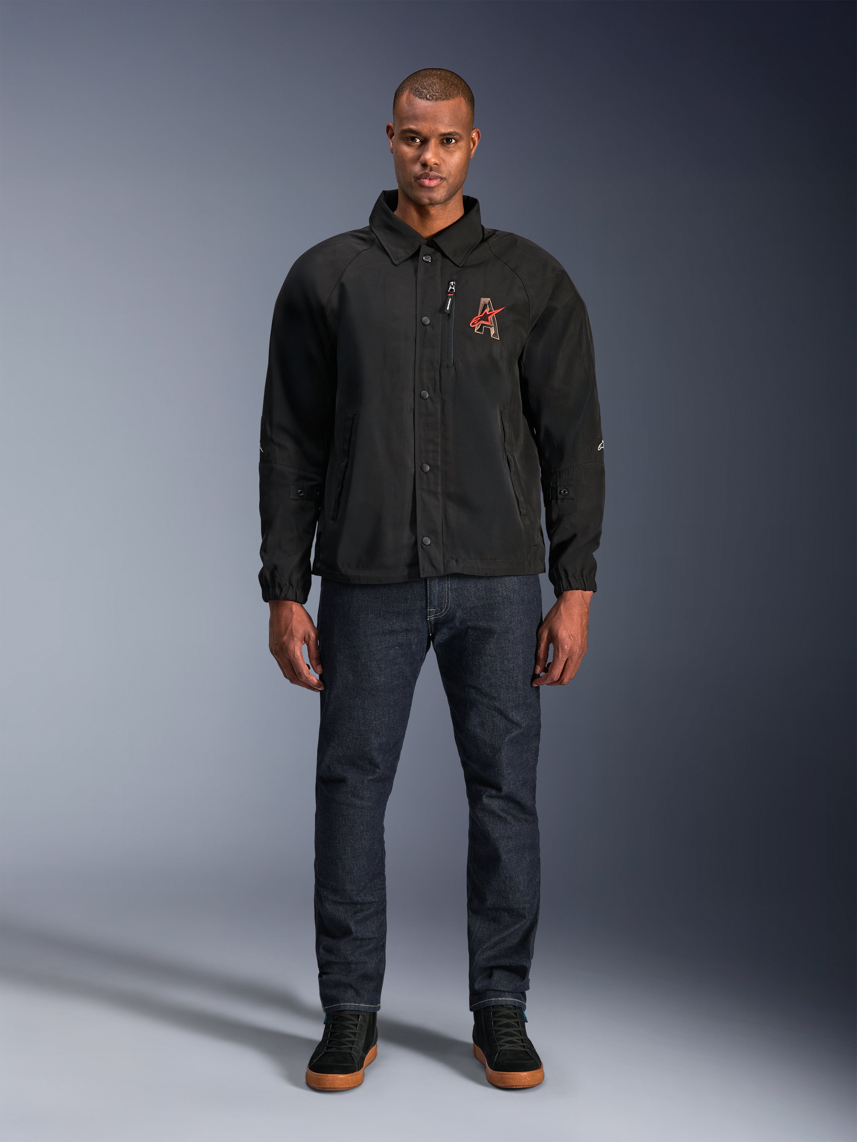 Alpinestars Revved WR Jas in het zwart, gedragen door een model met een donkerblauwe jeans en zwarte sneakers, vooraanzicht, met drukknopsluiting en een 3D geborduurd logo op de borst, urban contemporary motorkleding
