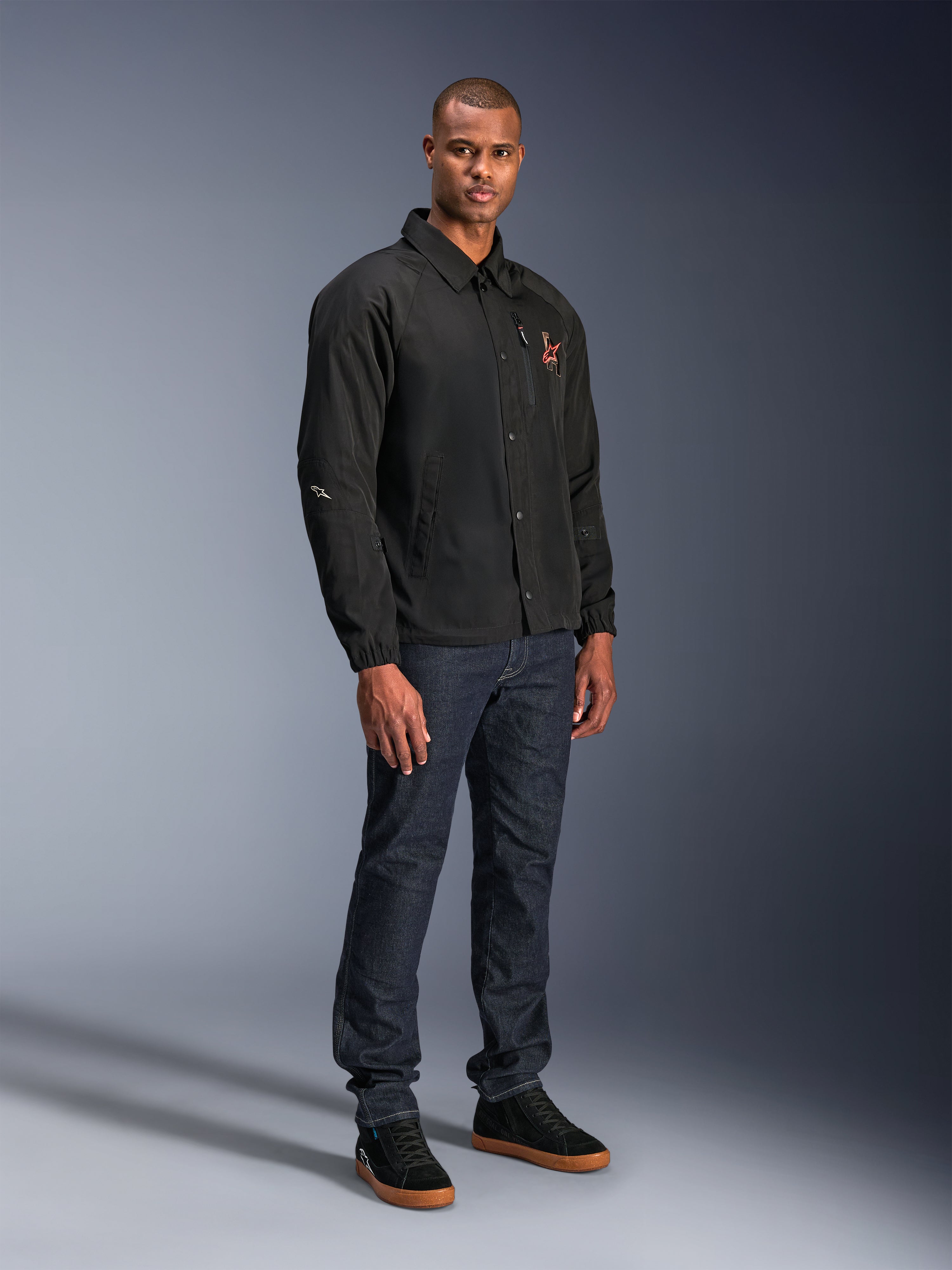 Alpinestars Revved WR Jacket in het zwart, urban motorjas in coaches-stijl gedragen door een model met donkerblauwe jeans en zwarte sneakers, voorzien van een drukknoopsluiting aan de voorzijde en een geborduurd logo, minimalistisch en sportief ontwerp voor rijden op de weg.