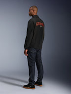 Alpinestars Revved WR Jas, zwart, met groot oranje 3D-geborduurd Alpinestars-logo op de rug, driekwart achteraanzicht van een model in donkerblauwe jeans en zwarte sneakers, urban contemporary motorjas met een coaches-jacket silhouet