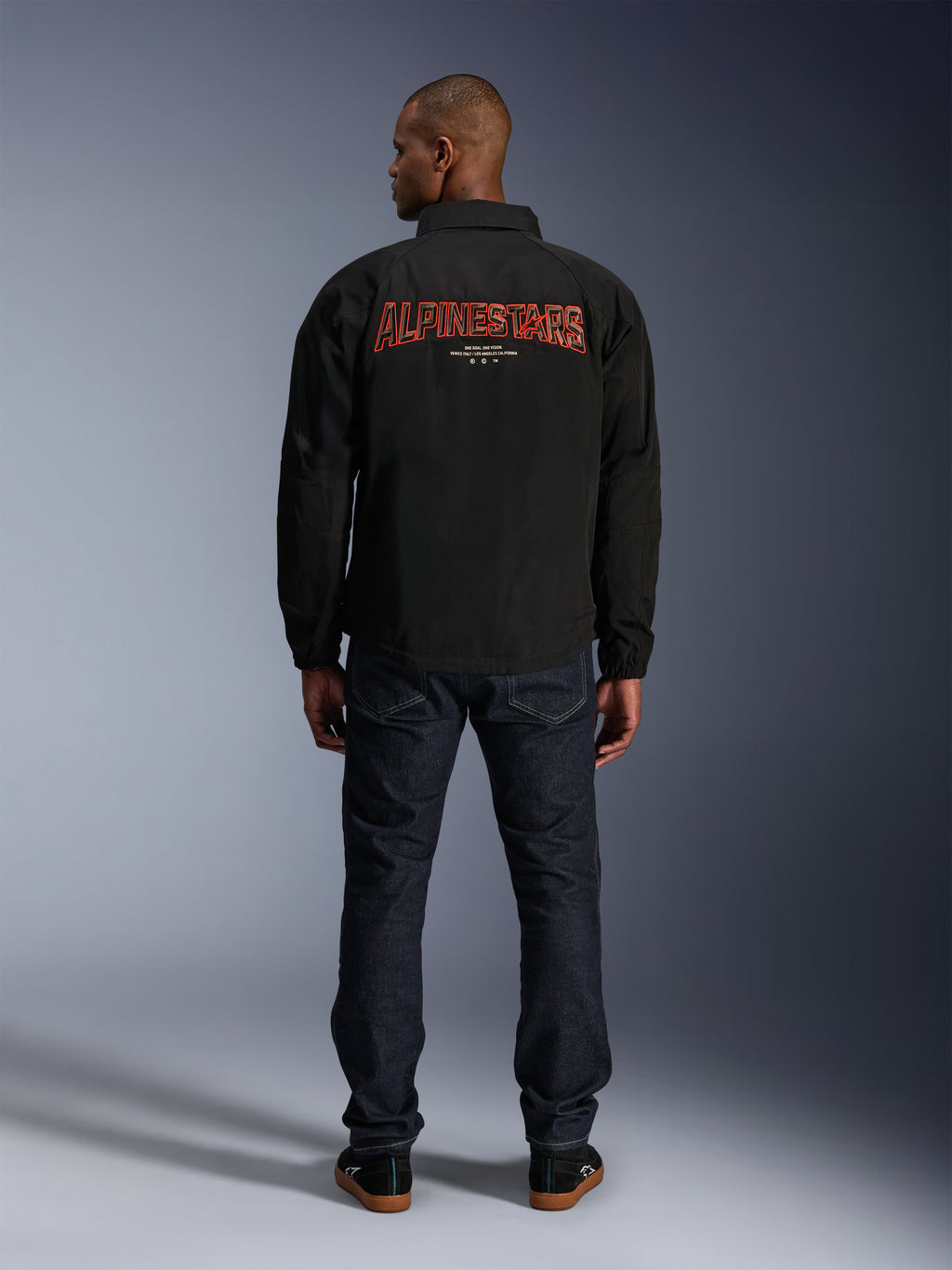 Alpinestars Revved WR Jas, zwart, achteraanzicht met groot rood 'ALPINESTARS' borduurwerk met 3D-effect, urban coachjack-stijl jas, gedragen met donkere denim jeans en zwarte sneakers, minimalistische motorkleding