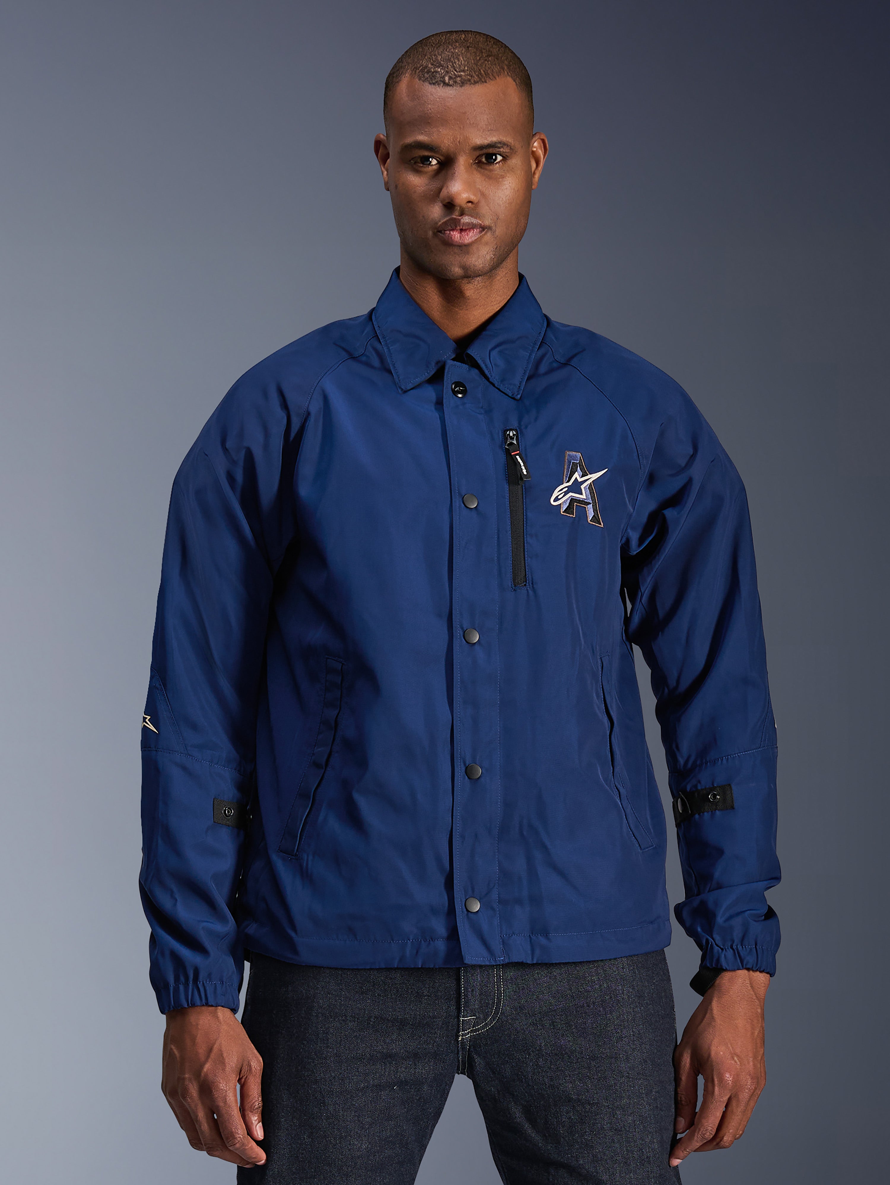 Alpinestars Revved WR Jacket, heren urban motorjas, navy blauw, coaches-stijl met omvouwbare kraag en drukknopen, zwarte borstzak met rits, groot 3D-geborduurd logo, verstelbare mouwlipjes, waterbestendig materiaal met satijnen afwerking, man draagt jas op donkere jeans