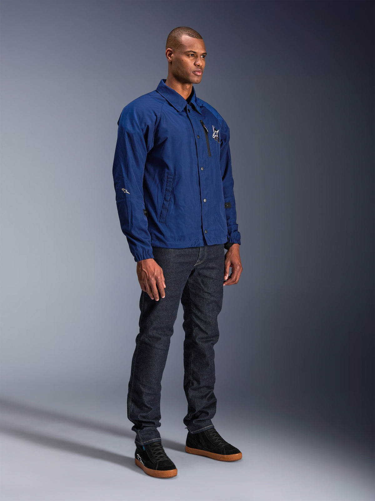 Alpinestars Revved WR Jacket, blauwe motorjas, gedragen door een mannelijk model met donkere denim jeans en zwarte sneakers, voorzien van een waterbestendig coaches jacket-silhouet met drukknoopsluiting en een minimalistisch logo op de borst.