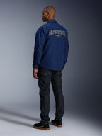 Model draagt de Alpinestars Revved WR Jas in marineblauw vanaf de achterkant gezien, voorzien van groot wit omlijnd 'ALPINESTARS' borduursel op de rug, gecombineerd met een donkere denim broek en zwarte sneakers, urban motor-esthetiek.