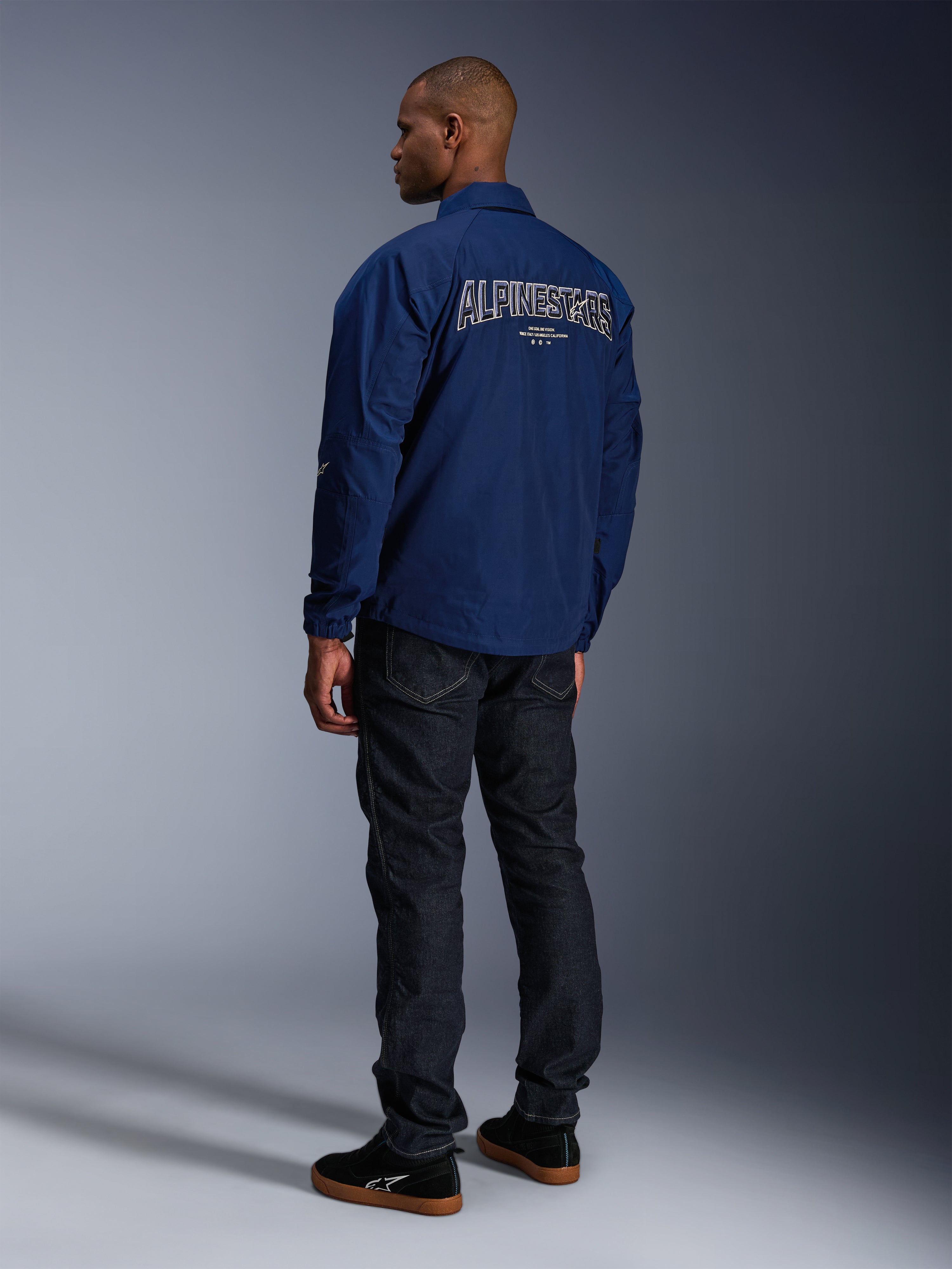Model draagt de Alpinestars Revved WR Jas in marineblauw vanaf de achterkant gezien, voorzien van groot wit omlijnd 'ALPINESTARS' borduursel op de rug, gecombineerd met een donkere denim broek en zwarte sneakers, urban motor-esthetiek.