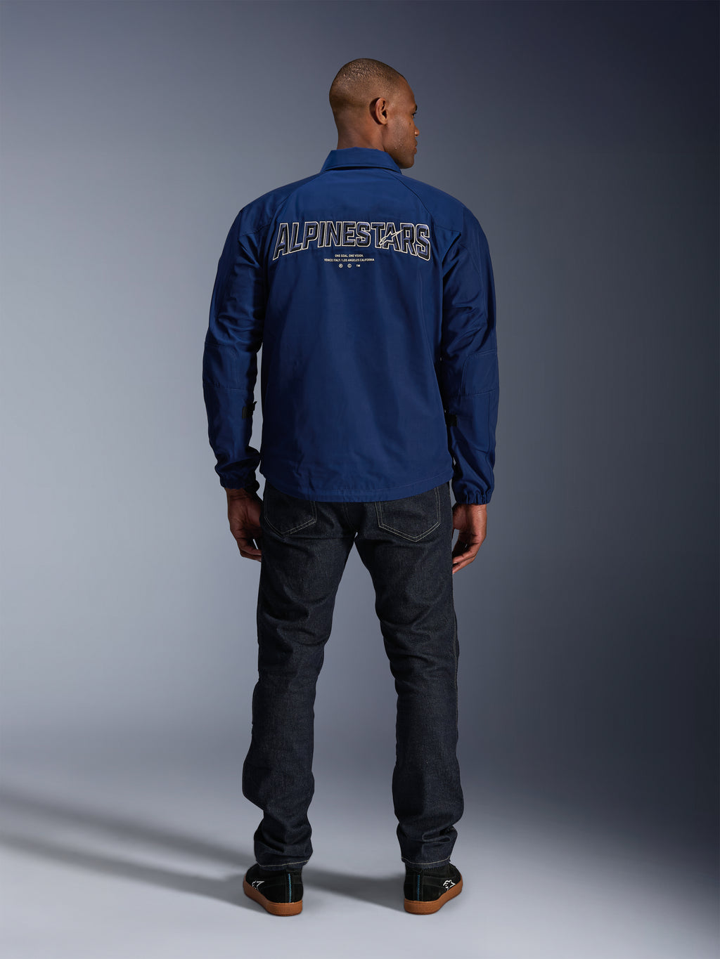 Alpinestars Revved WR jas in donkerblauw, achteraanzicht, met groot 'ALPINESTARS' borduursel met 3D-effect op de rug, gestyled met een donkere denim jeans en zwarte sneakers, urban motorkleding.