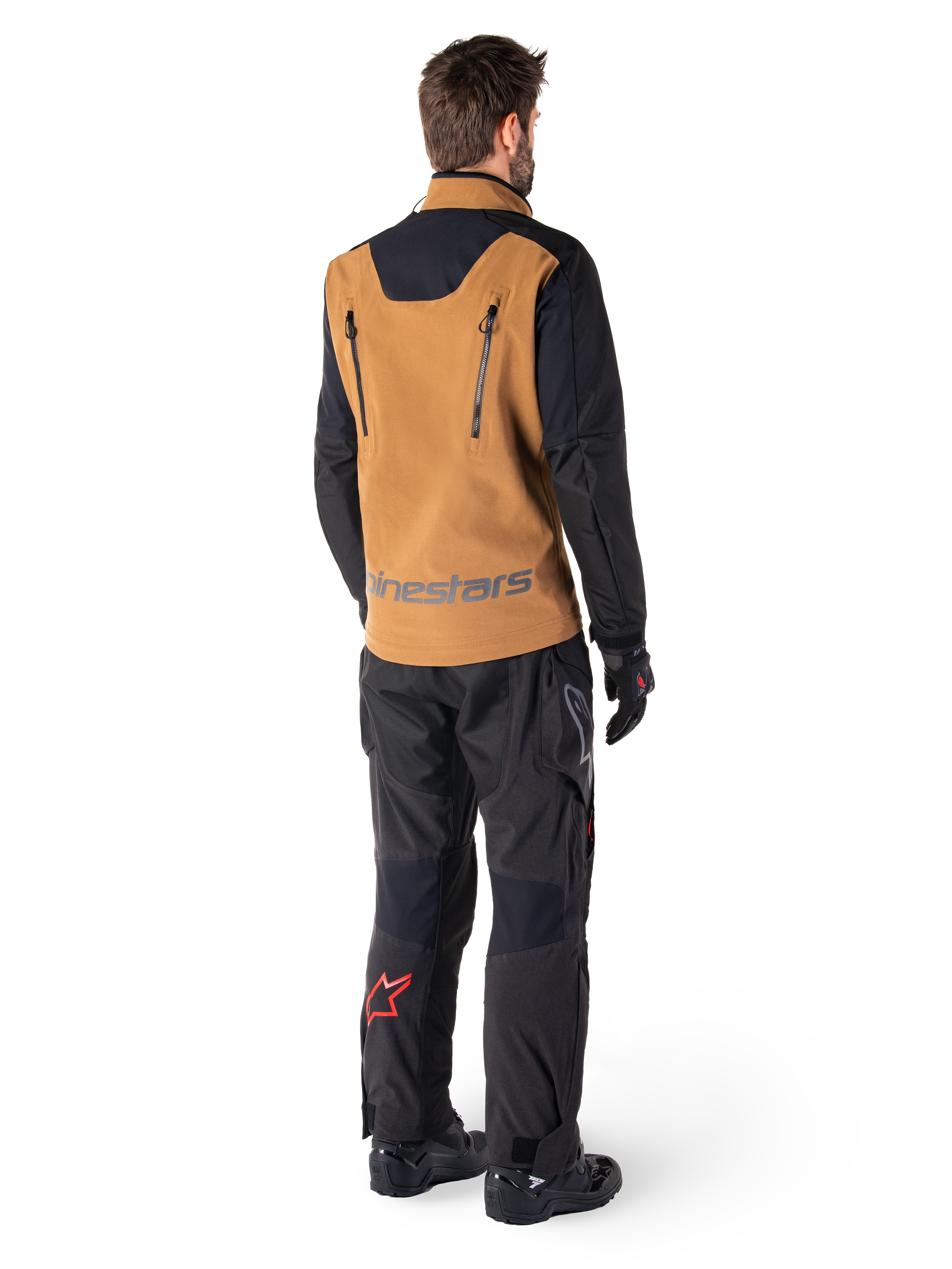 Alpinestars Hyde XT Stretch Drystar®XF jas en Hyde XT Drystar® broek, achteraanzicht van een rijder in zandkleurige en zwarte adventure touring uitrusting, jas met verticale rugritsen en Alpinestars-branding, zwarte broek met rood sterlogo op de kuit, gecombineerd met zwarte handschoenen en laarzen.