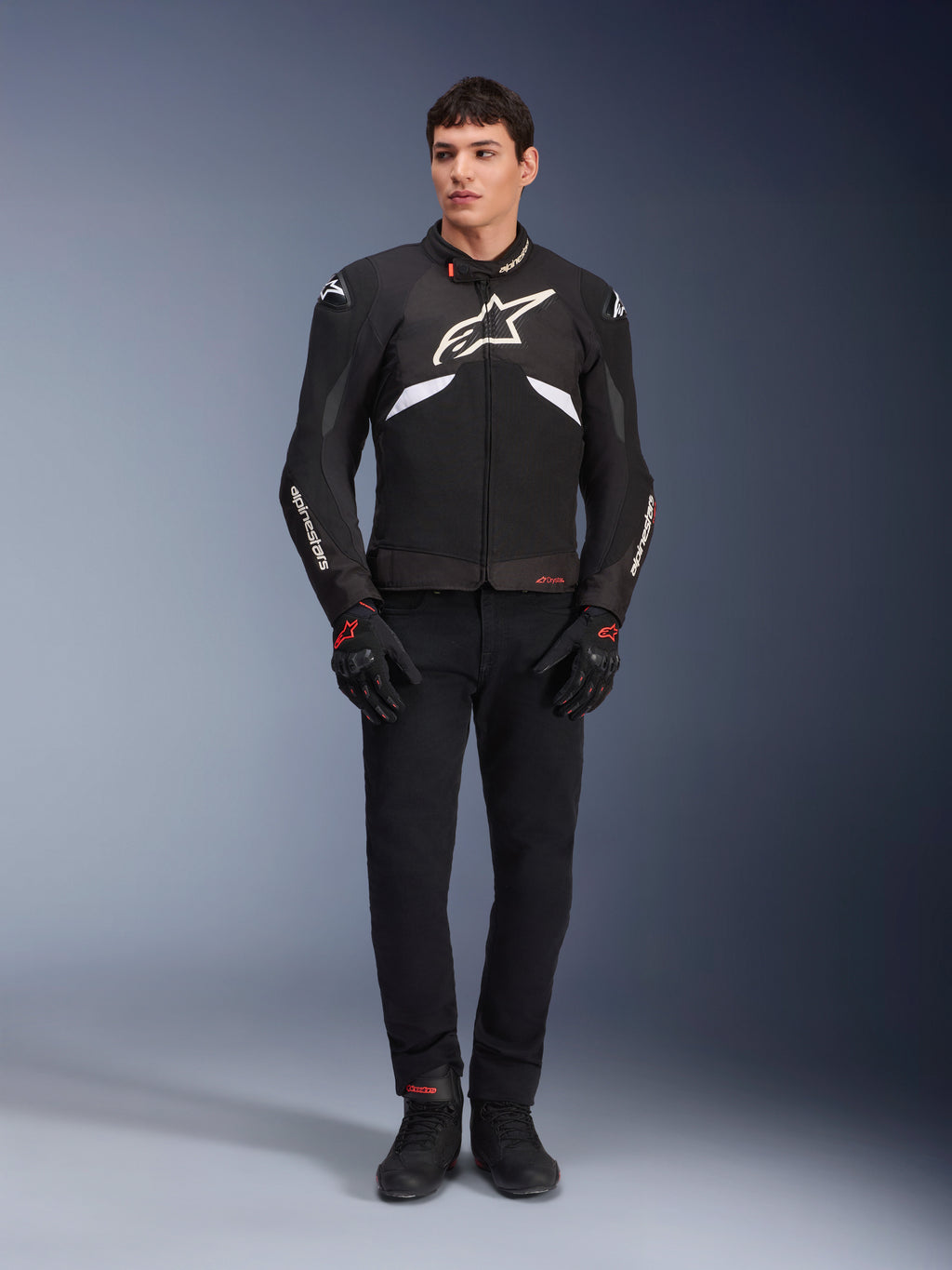 Mannelijk model draagt de Alpinestars T-GP R V3 Drystar® Jas in zwart en wit, waterdichte textiele sportjas met groot wit Alpinestars-logo op de borst, gedragen met een zwarte motorbroek en handschoenen, professioneel studioportret dat het slanke sportieve silhouet benadrukt.