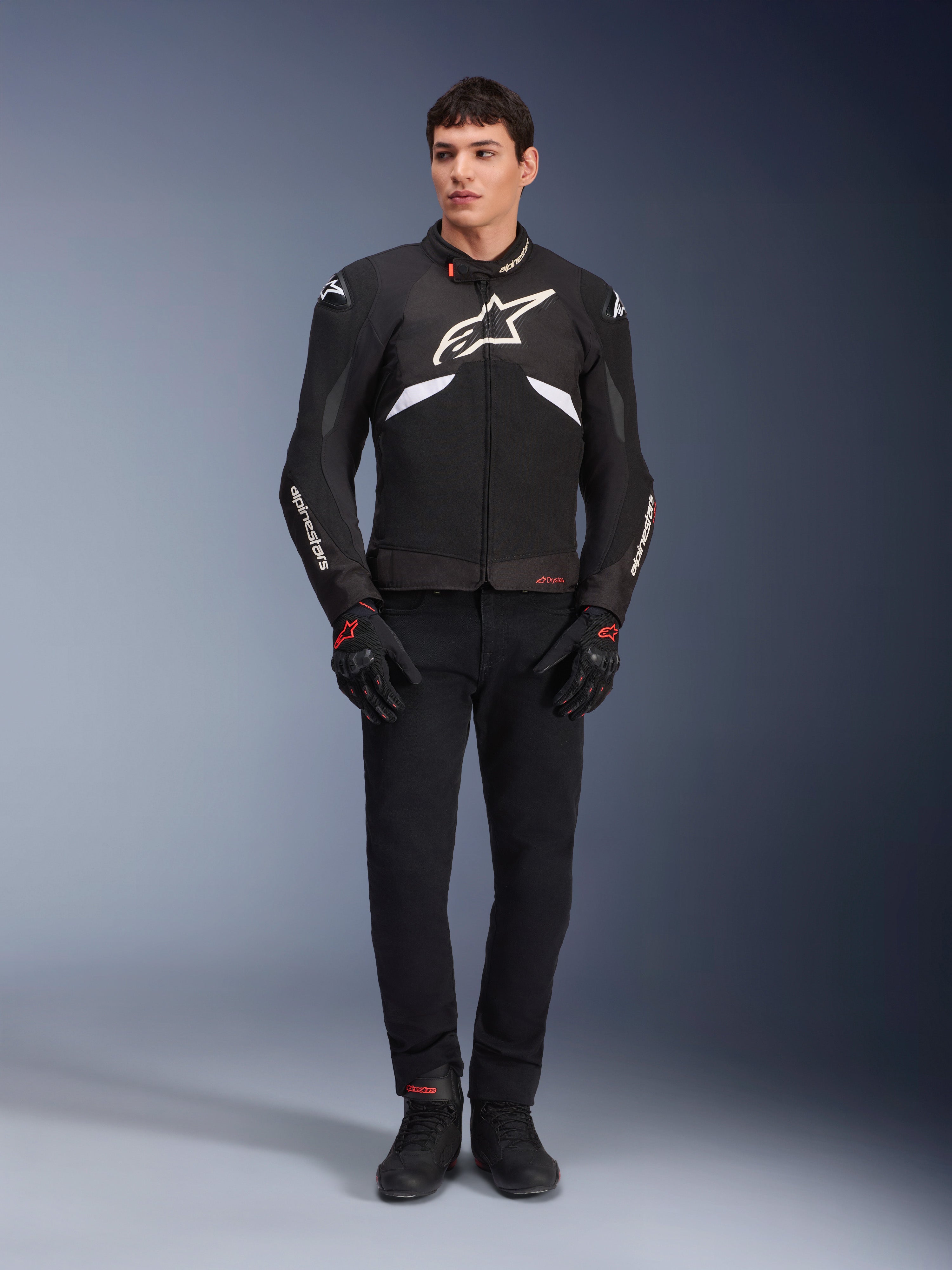 Mannelijk model draagt de Alpinestars T-GP R V3 Drystar® Jas in zwart en wit, waterdichte textiele sportjas met groot wit Alpinestars-logo op de borst, gedragen met een zwarte motorbroek en handschoenen, professioneel studioportret dat het slanke sportieve silhouet benadrukt.