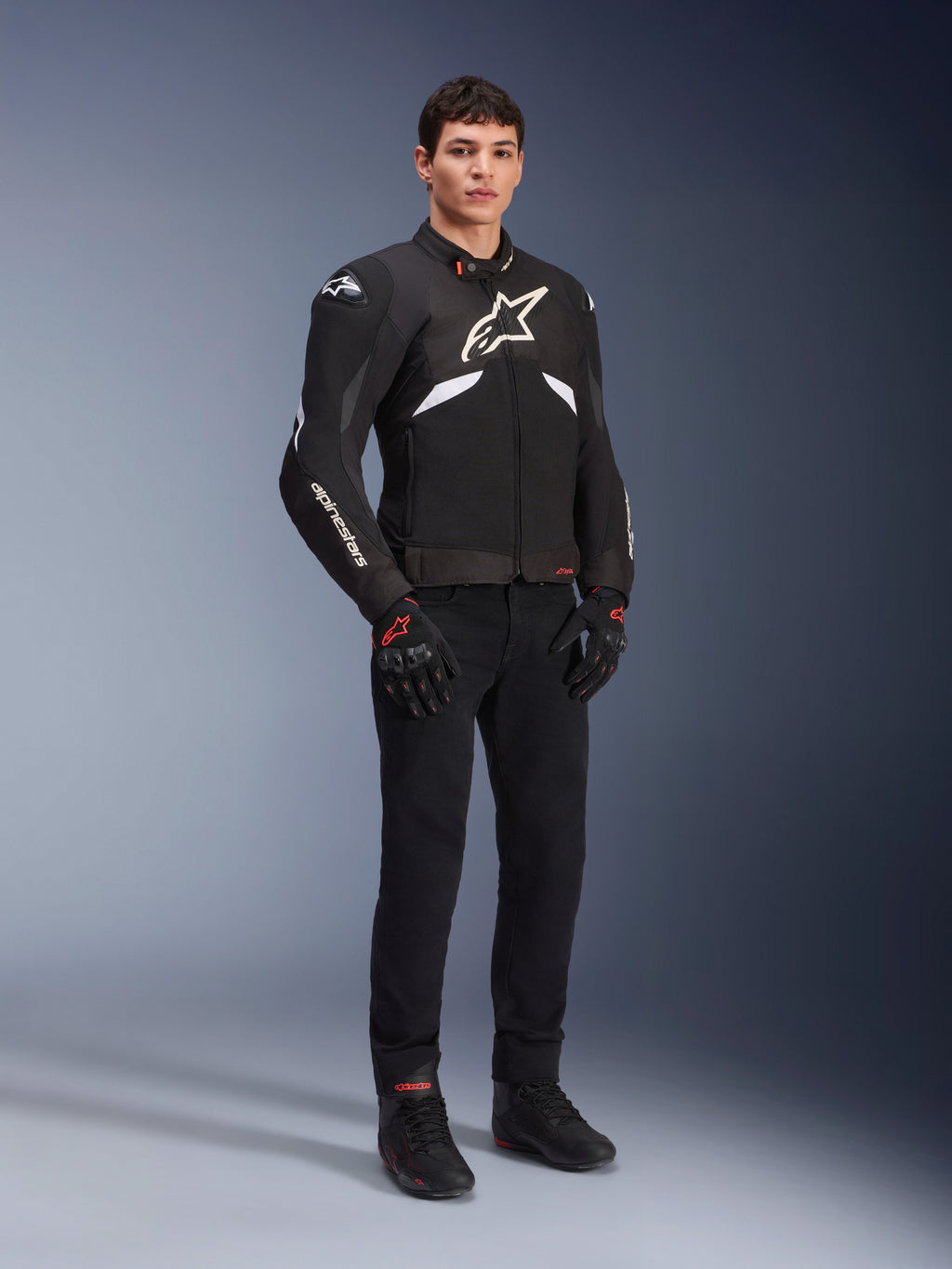 Alpinestars T-GP R V3 Drystar® Jas in zwart en wit, gedragen door een model met zwarte motorhandschoenen en broek, met een wit logo op de borst, witte mouwaccenten en beschermende schoudersliders, waterdichte sportjas van textiel voor op de weg