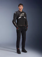 Alpinestars T-GP R V3 Drystar® Jas in zwart en wit, gedragen door een model met zwarte motorhandschoenen en broek, met een wit logo op de borst, witte mouwaccenten en beschermende schoudersliders, waterdichte sportjas van textiel voor op de weg