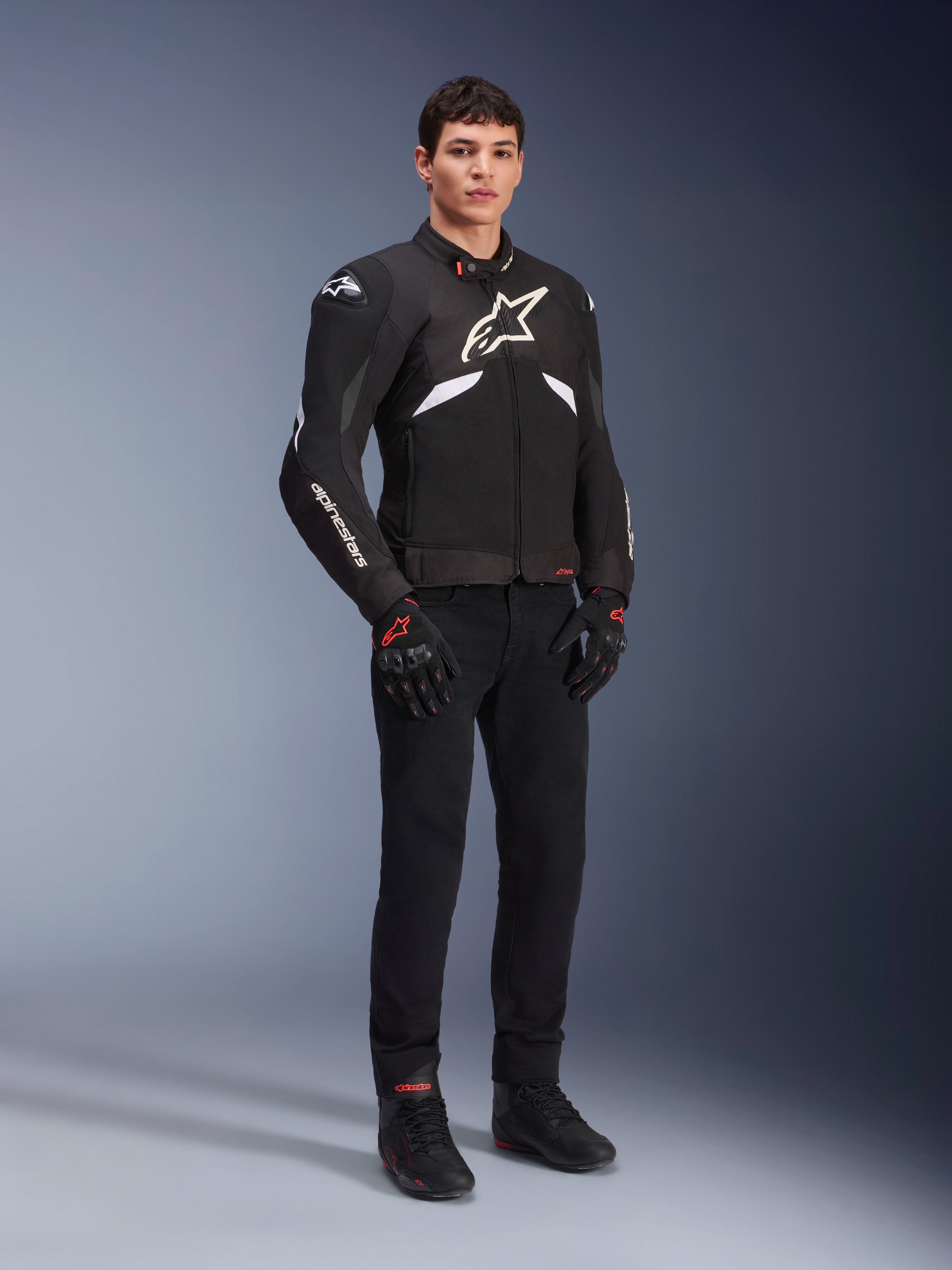 Alpinestars T-GP R V3 Drystar® Jas in zwart en wit, gedragen door een model met zwarte motorhandschoenen en broek, met een wit logo op de borst, witte mouwaccenten en beschermende schoudersliders, waterdichte sportjas van textiel voor op de weg