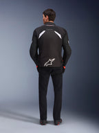 Alpinestars T-GP R V3 Drystar® Jas, zwart en wit, achteraanzicht, waterdichte textiele motorjas met een groot wit sterlogo op de onderrug en witte accenten op de schouders, gecombineerd met een zwarte broek en laarzen, race-afgeleid sportief ontwerp.