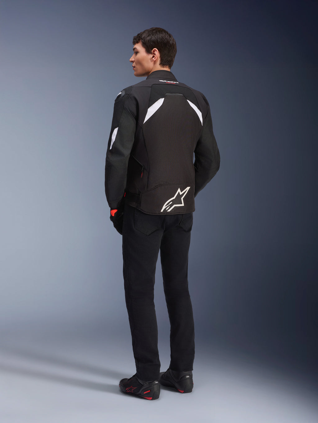 Alpinestars T-GP R V3 Drystar® Jas in zwart en wit, sportieve textiele motorjas, mannelijk model van de achterkant, witte schouderaccenten, groot wit logo op de onderrug, gecombineerd met zwarte motorbroek en laarzen.