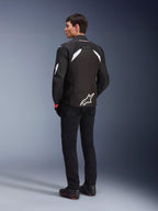 Alpinestars T-GP R V3 Drystar® Jas in zwart en wit, sportieve textiele motorjas, mannelijk model van de achterkant, witte schouderaccenten, groot wit logo op de onderrug, gecombineerd met zwarte motorbroek en laarzen.