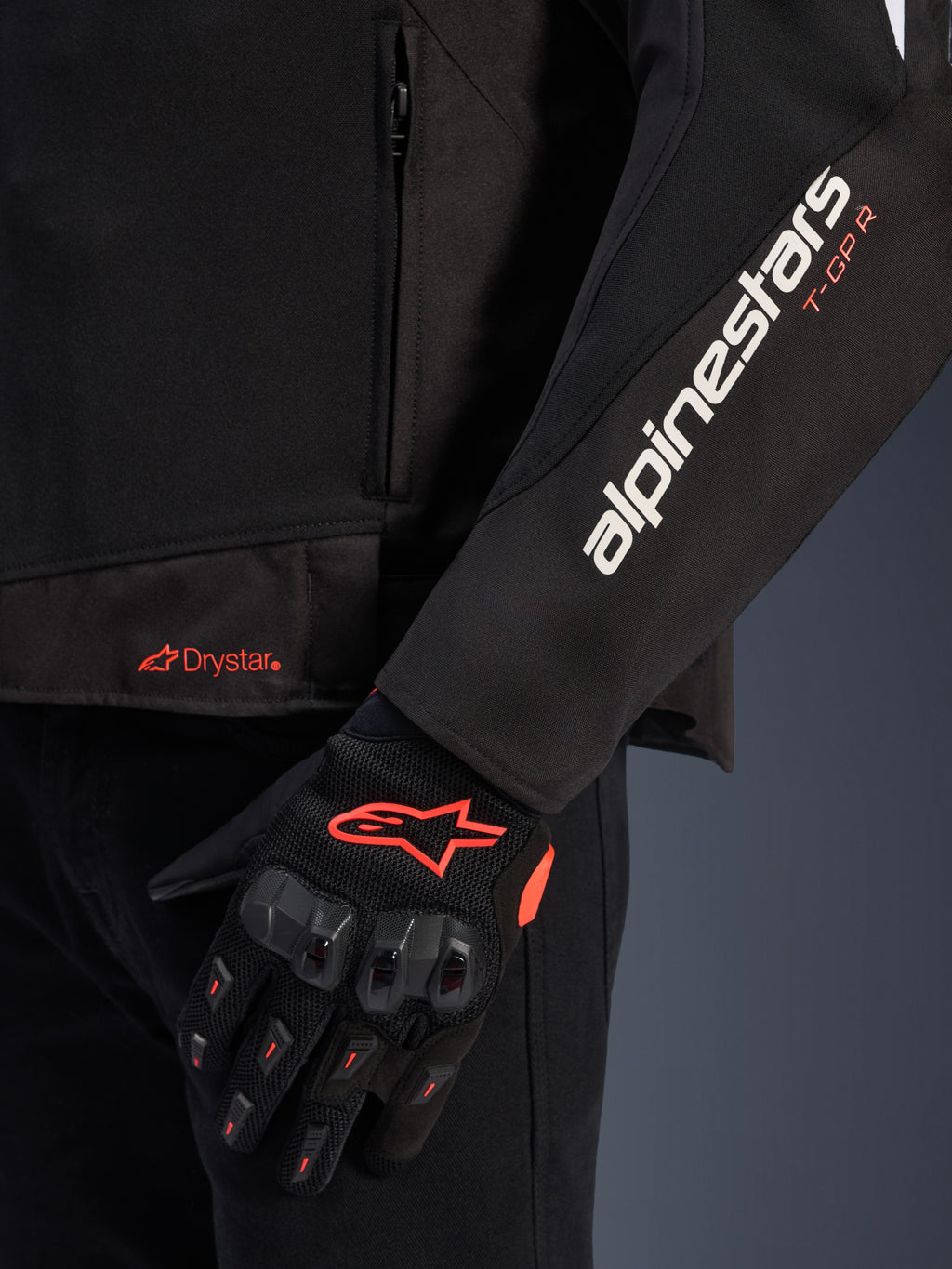 Alpinestars T-GP R V3 Drystar® Jacket, zwarte textiele motorjas, close-up met witte Alpinestars- en oranje T-GP R-branding op de mouw, oranje Drystar-logo op de zoom, gecombineerd met zwarte beschermende mesh handschoenen met rode accenten.