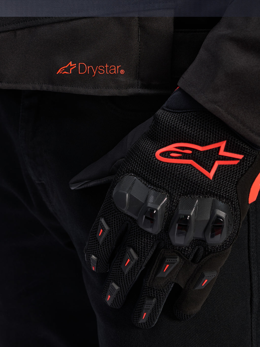 Close-up van de taille van de Alpinestars T-GP R V3 Drystar® jas met een rood Drystar-logo, getoond met een zwarte motorhandschoen met een rood Alpinestars-sterlogo, mesh-stof en versterkte knokkelbescherming.