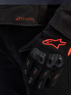 Close-up van de taille van de Alpinestars T-GP R V3 Drystar® jas met een rood Drystar-logo, getoond met een zwarte motorhandschoen met een rood Alpinestars-sterlogo, mesh-stof en versterkte knokkelbescherming.