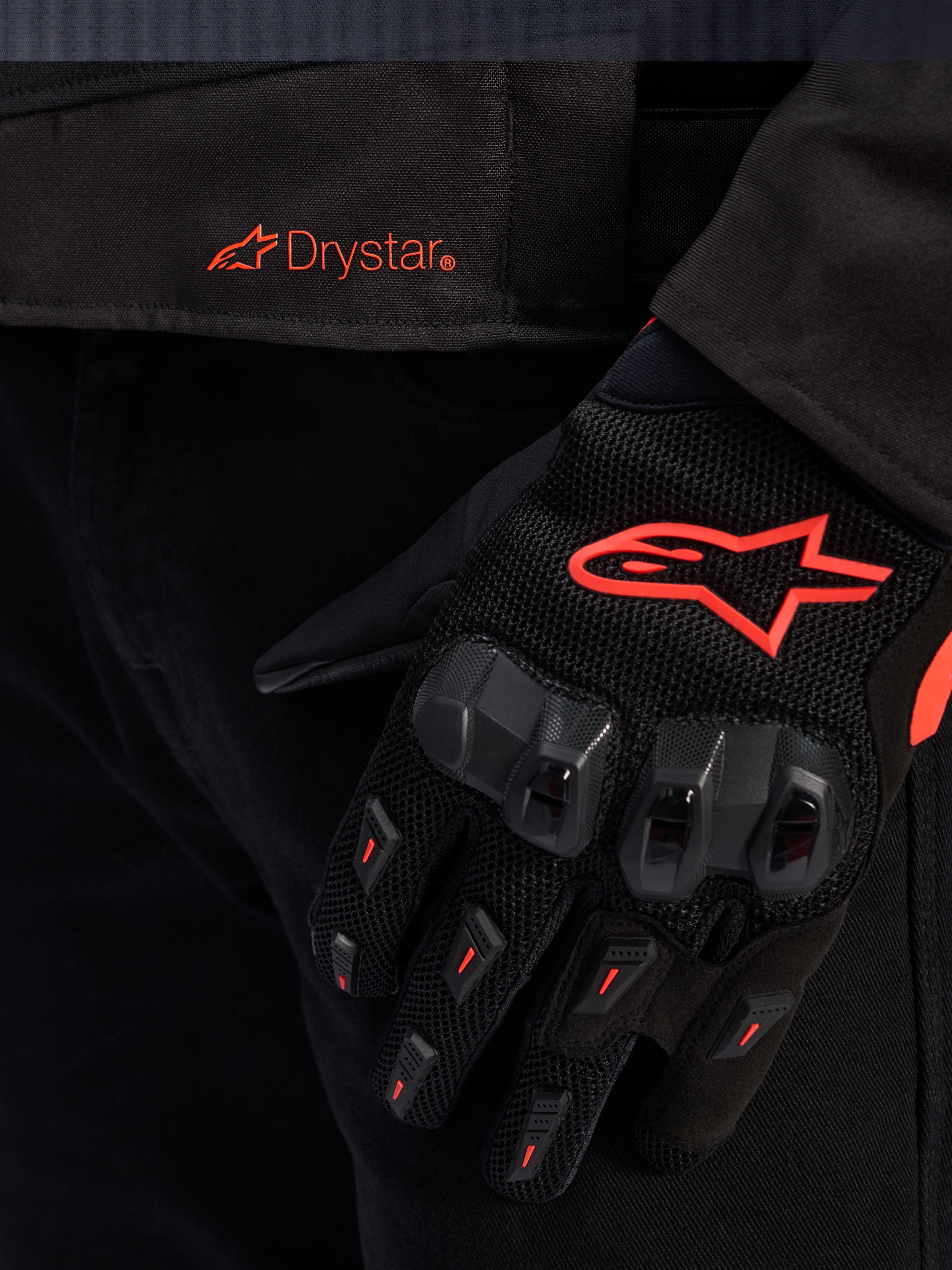 Close-up van de taille van de Alpinestars T-GP R V3 Drystar® jas met een rood Drystar-logo, getoond met een zwarte motorhandschoen met een rood Alpinestars-sterlogo, mesh-stof en versterkte knokkelbescherming.