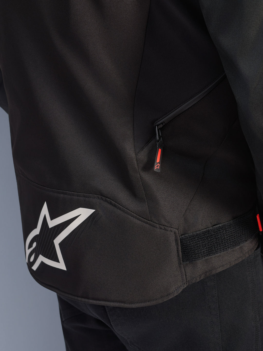 Alpinestars T-GP R V3 Drystar® Jacket, zwart en wit, detailopname van de onderrug met een groot wit Alpinestars sterlogo, een zijzak met rits en rood lipje, en een klittenband verstelband in de taille, gemaakt van duurzaam textiel.