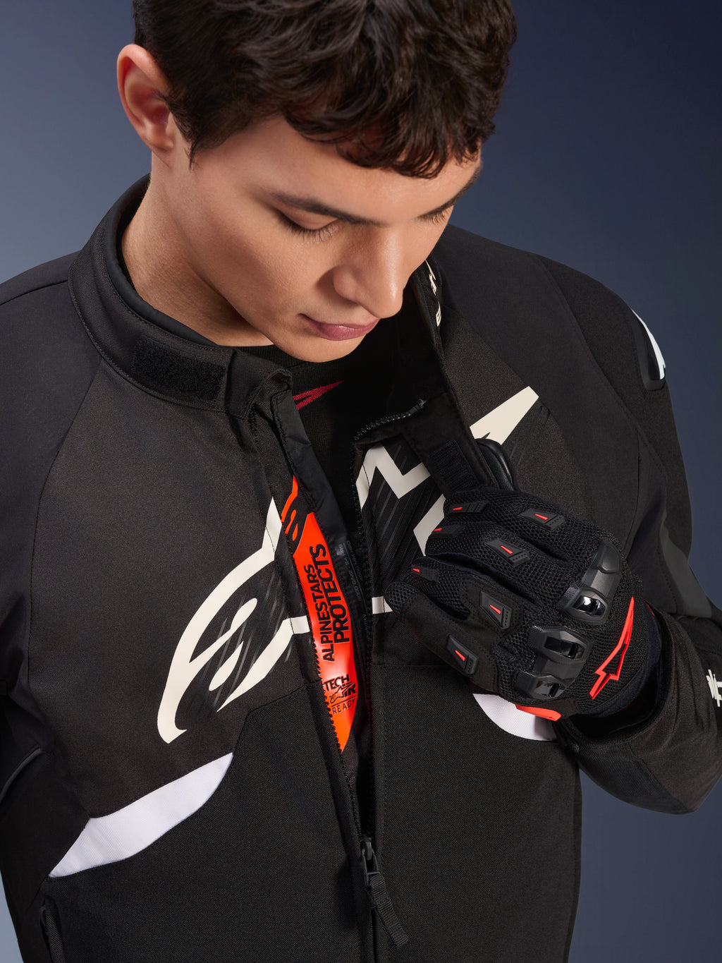 Alpinestars T-GP R V3 Drystar® Jas, zwart-witte sportieve motorjas van textiel, gedeeltelijk opengeritst met zichtbare oranje 'Alpinestars Protects' binnenband, gedragen met zwart-rode mesh handschoenen, race-afgeleide beschermende kleding