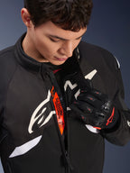 Alpinestars T-GP R V3 Drystar® Jas, zwart-witte sportieve motorjas van textiel, gedeeltelijk opengeritst met zichtbare oranje 'Alpinestars Protects' binnenband, gedragen met zwart-rode mesh handschoenen, race-afgeleide beschermende kleding