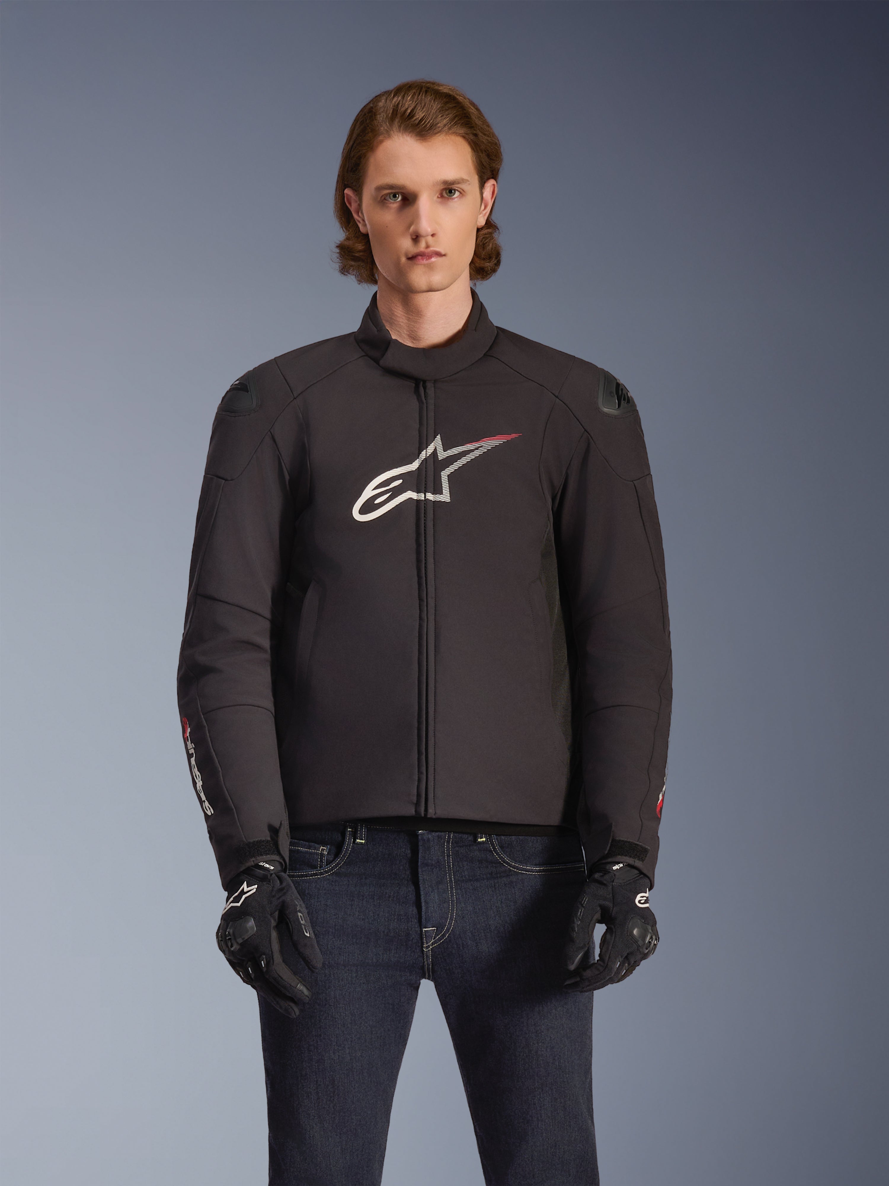 Alpinestars SMX Waterproof Jacket, zwarte textielen motorjas met wit en rood sterlogo op de borst, gedragen door een model met zwarte handschoenen en blauwe jeans, voorzien van externe schoudersliders en een sportieve pasvorm.