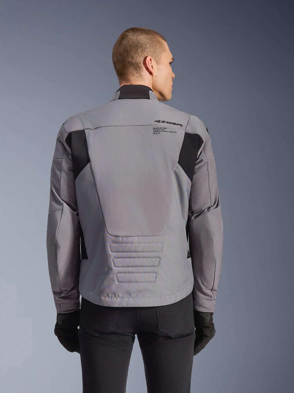 Alpinestars SMX Waterproof jas, grijs met zwarte accenten, achteraanzicht met branding op de bovenrug en verlengd achterprofiel, gedragen door een model met zwarte broek en handschoenen