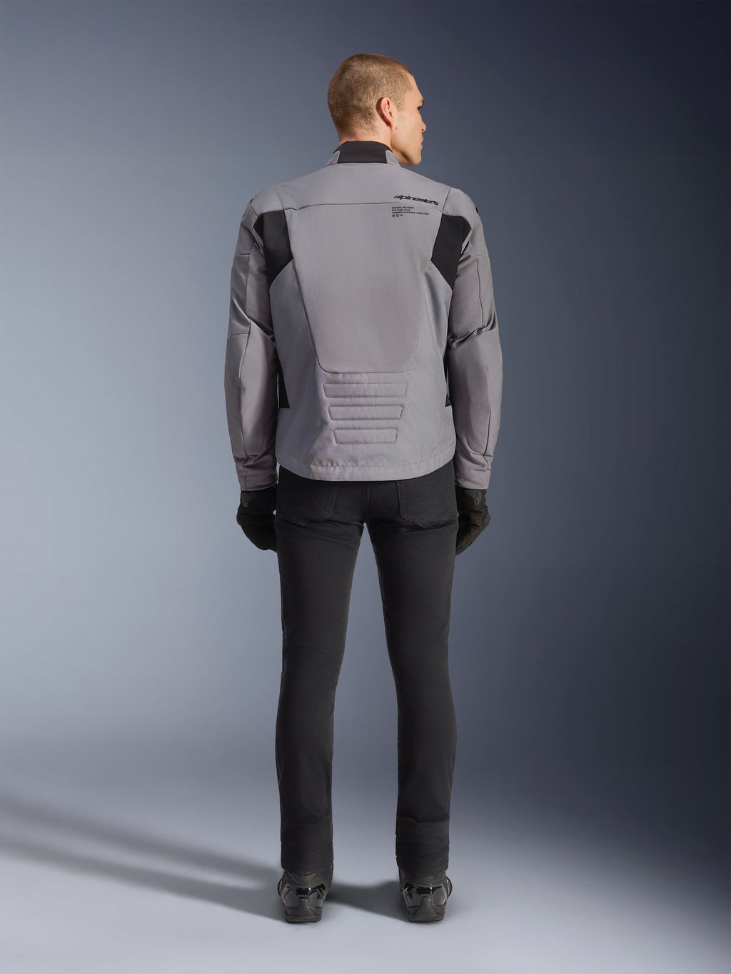 Alpinestars SMX Waterproof Jacket, achteraanzicht, donkergrijs en zwart, voorzien van zwarte textielpanelen op de schouders en een sportieve pasvorm, gecombineerd met een zwarte broek en handschoenen voor een complete motorlook