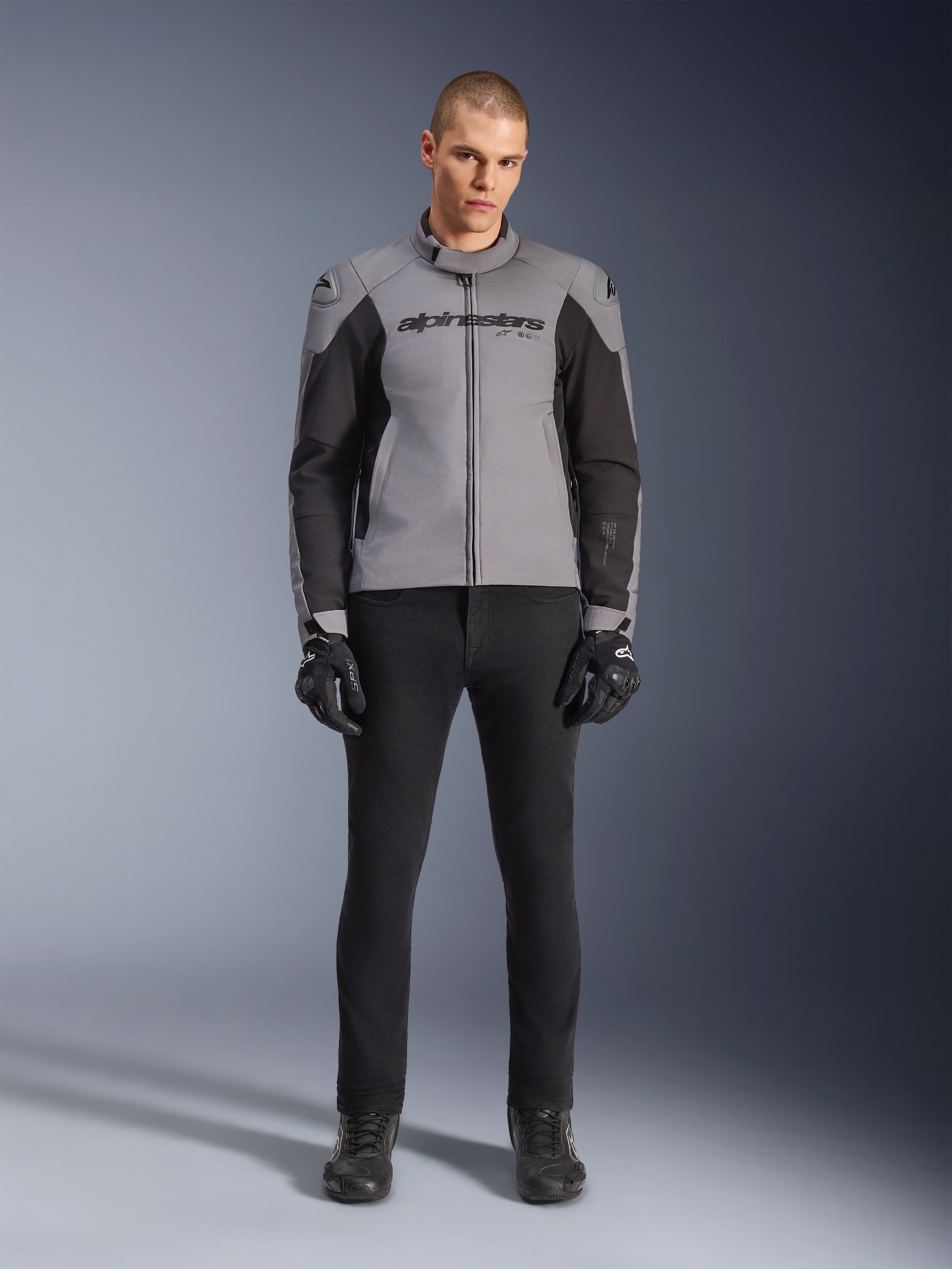 Alpinestars SMX Waterproof Jas, donkergrijs en zwart, vooraanzicht gedragen door een mannelijk model, textiele sportieve motorjas met zwarte zijpanelen en Alpinestars-branding op de borst, gecombineerd met een zwarte motorbroek en handschoenen.